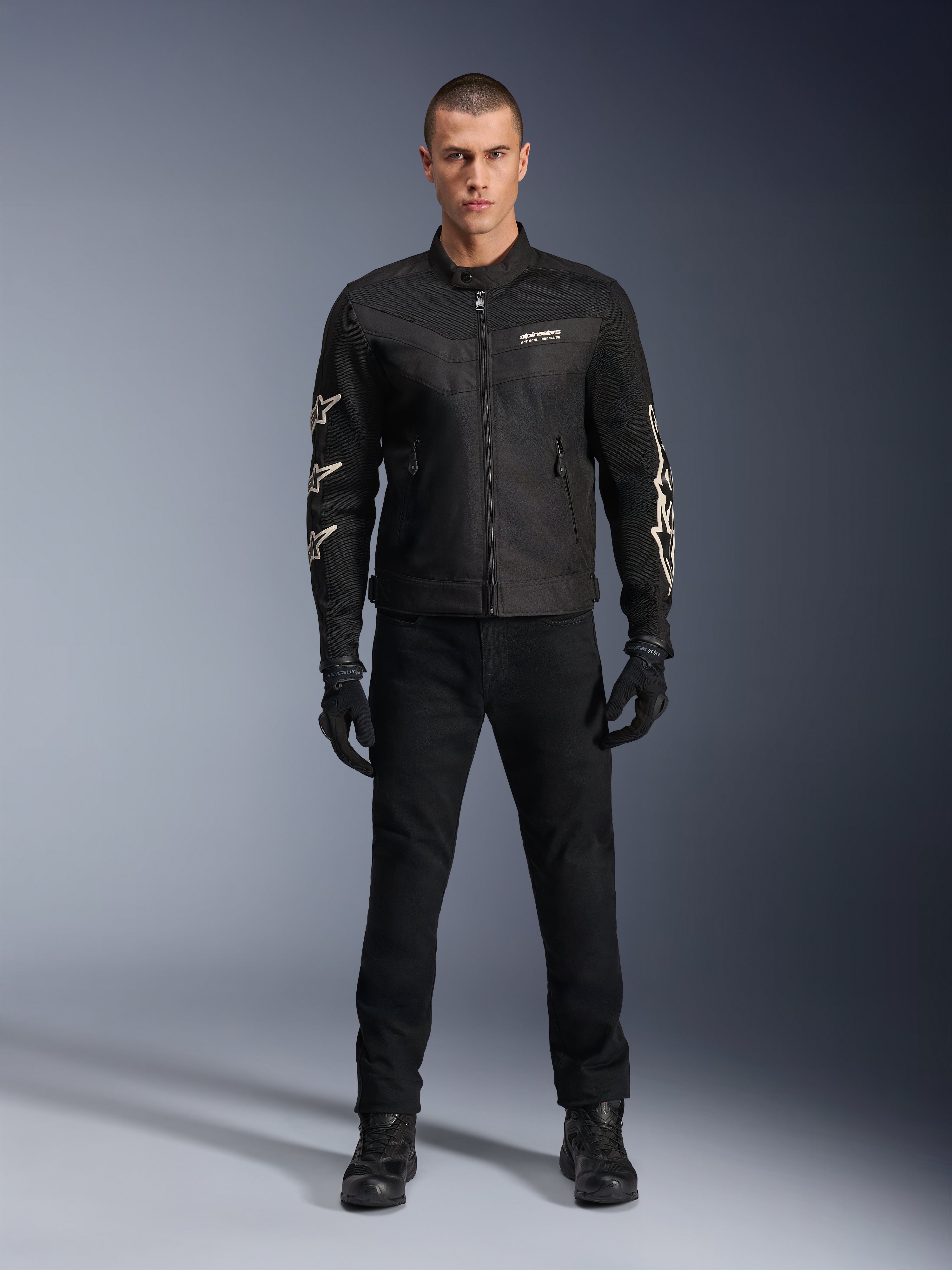 Model w kurtce Alpinestars T-Dyno Air, czarna, lekka motocyklowa kurtka tekstylna z oddychającą siatką 3D, białe logo gwiazdy na rękawach, zestawiona z czarnymi spodniami, rękawicami i butami, widok z przodu