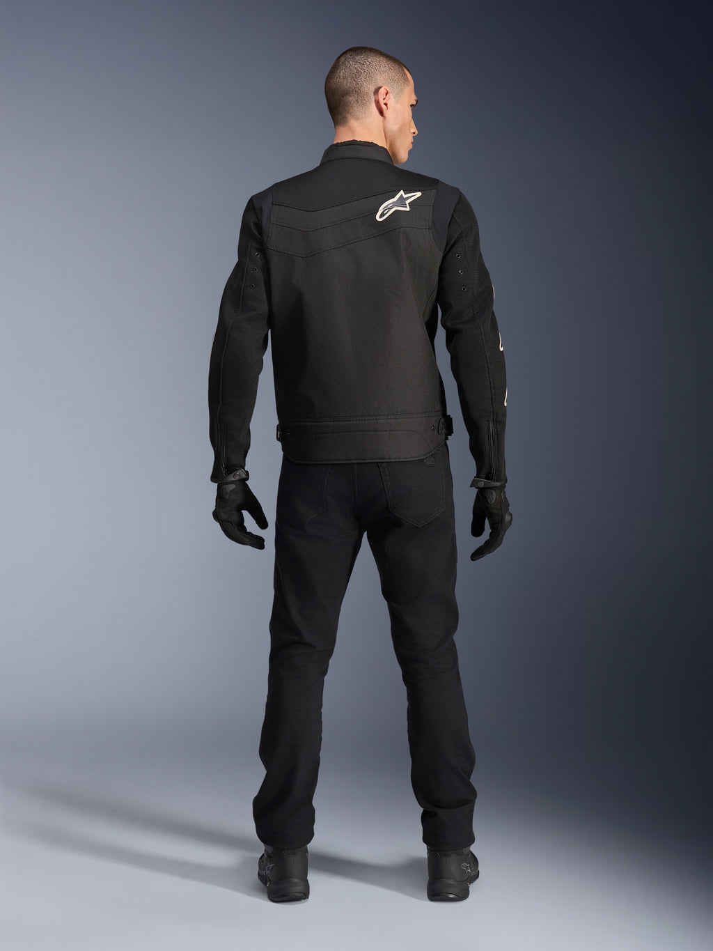 Alpinestars T-Dyno Air Jacket, czarna, widok z tyłu na modelu, tekstylna kurtka motocyklowa z panelami z siatki 3D i białym logo gwiazdy na ramieniu, noszona z czarnymi spodniami i rękawicami, nowoczesny miejski strój motocyklowy
