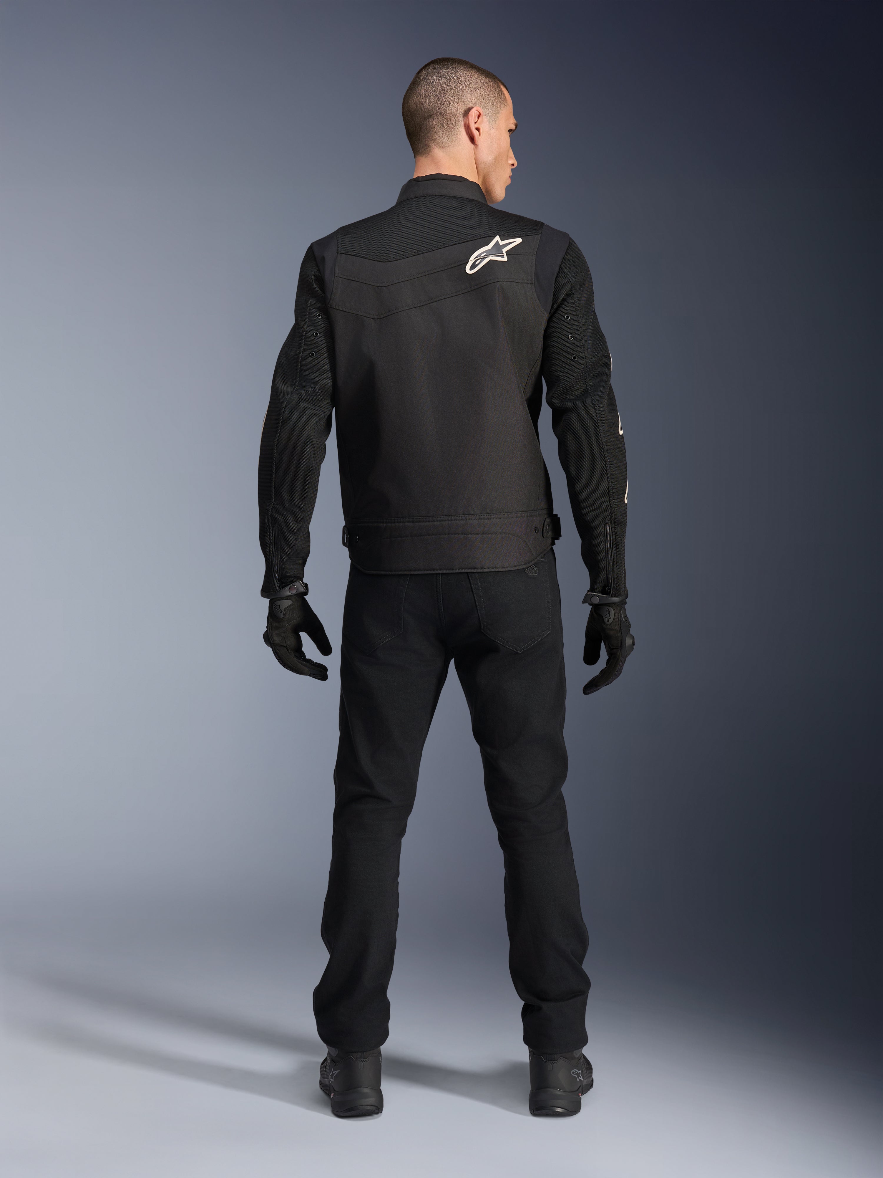 Alpinestars T-Dyno Air Jacket, czarna, widok z tyłu na modelu, tekstylna kurtka motocyklowa z panelami z siatki 3D i białym logo gwiazdy na ramieniu, noszona z czarnymi spodniami i rękawicami, nowoczesny miejski strój motocyklowy