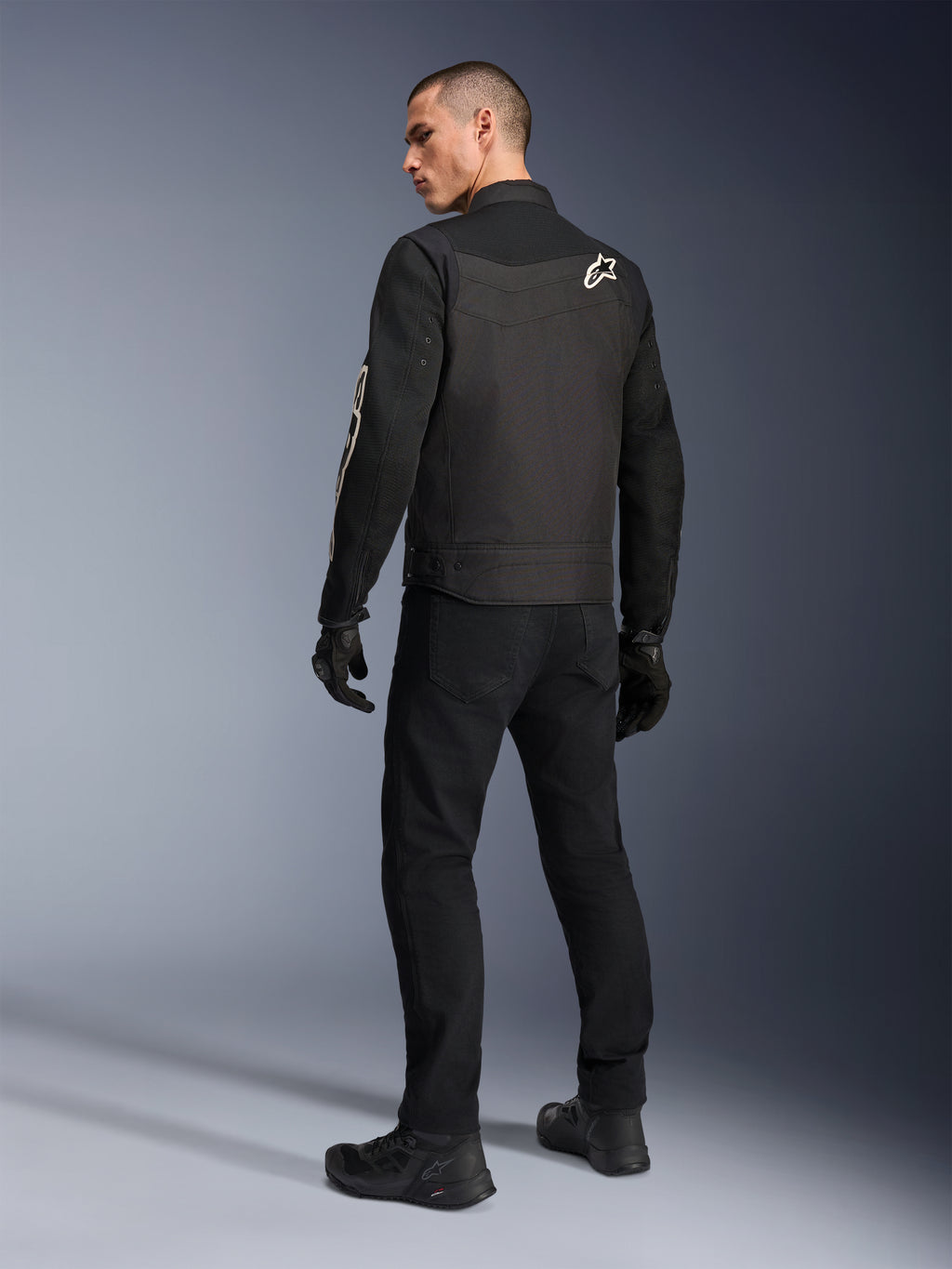 Alpinestars T-Dyno Air Jacket, czarna, widok od tyłu modelu w oddychającej tekstylnej kurtce motocyklowej, z białym logo Alpinestars na ramieniu, w zestawie z czarnymi spodniami i rękawicami, miejski styl jazdy