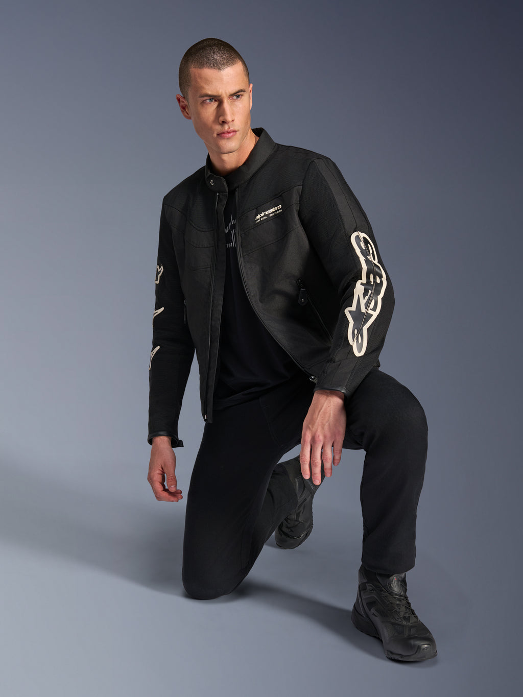 Męski model w kurtce Alpinestars T-Dyno Air, czarna tekstylna kurtka motocyklowa z dużym białym logo na rękawach, w zestawieniu z czarnymi spodniami i butami motocyklowymi, nowoczesny design inspirowany stylem vintage z panelami z siatki 3D.