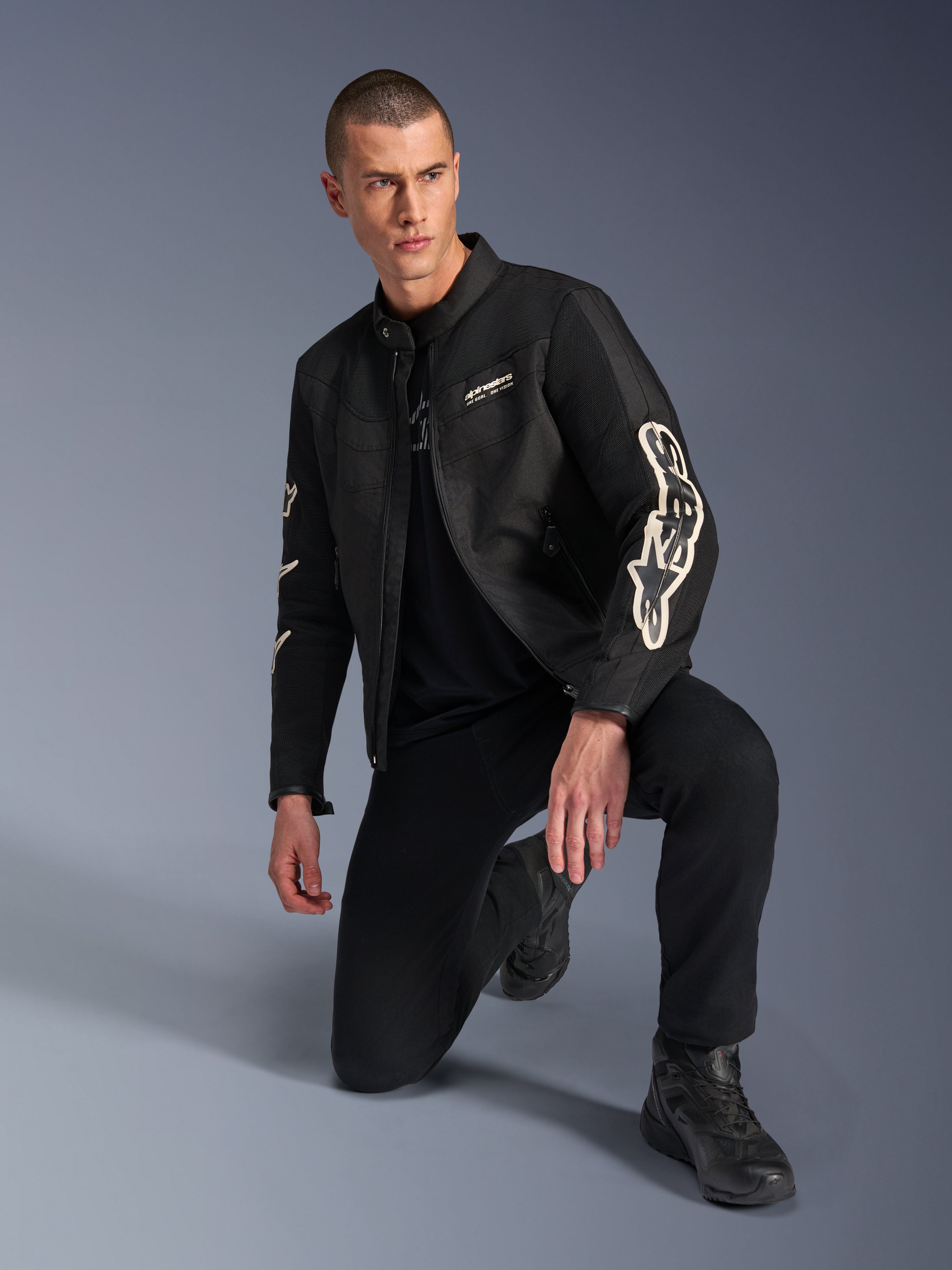 Męski model w kurtce Alpinestars T-Dyno Air, czarna tekstylna kurtka motocyklowa z dużym białym logo na rękawach, w zestawieniu z czarnymi spodniami i butami motocyklowymi, nowoczesny design inspirowany stylem vintage z panelami z siatki 3D.