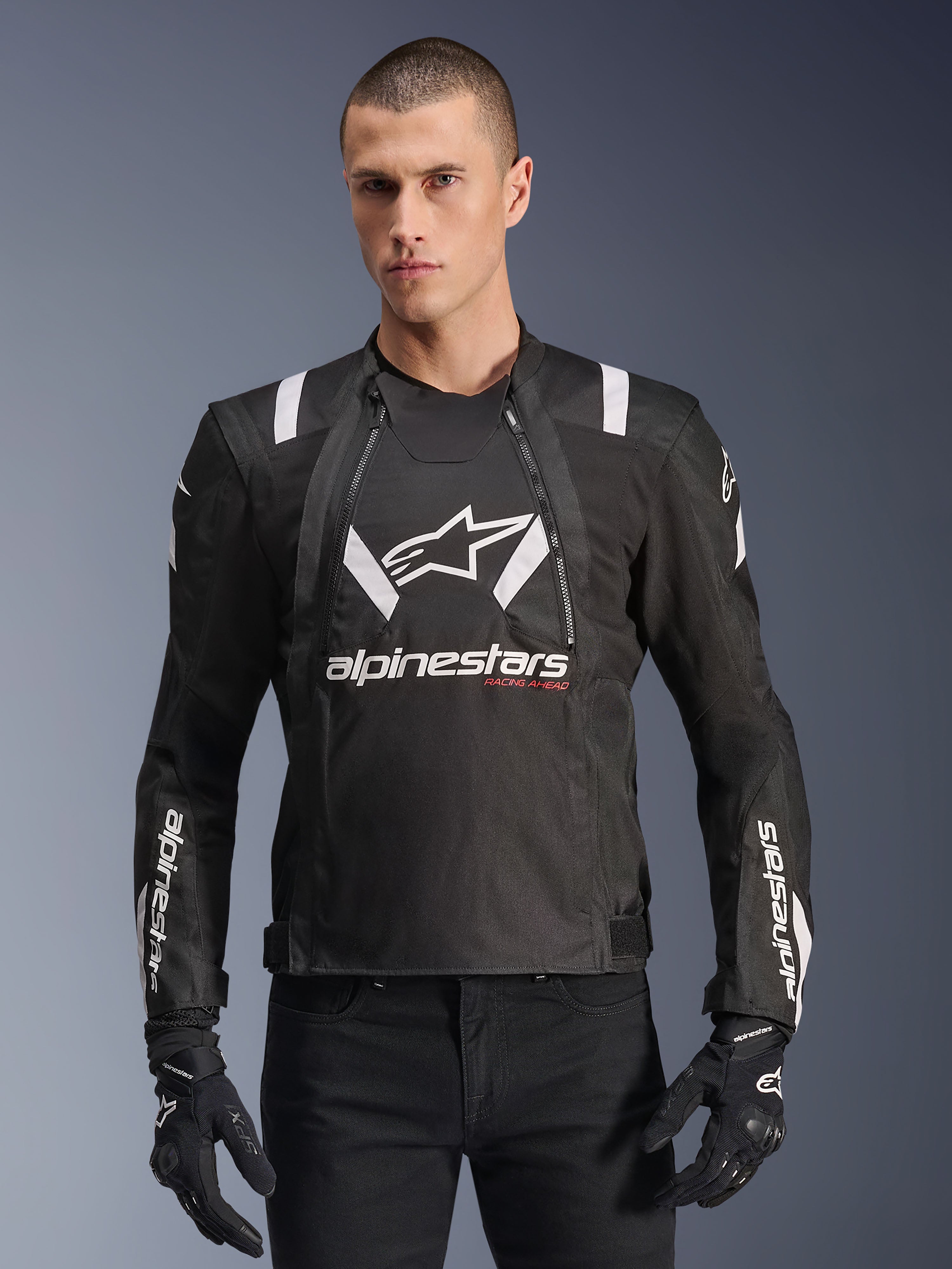 Alpinestars T-Stunt Air Jacket, czarno-biała kurtka motocyklowa, noszona przez mężczyznę w czarnych rękawicach i spodniach, z wyrazistym białym logo na klatce piersiowej i rękawach oraz czerwonymi akcentami „Racing Ahead”, sportowy design z białymi paskami na ramionach i oddychającymi panelami z siatki