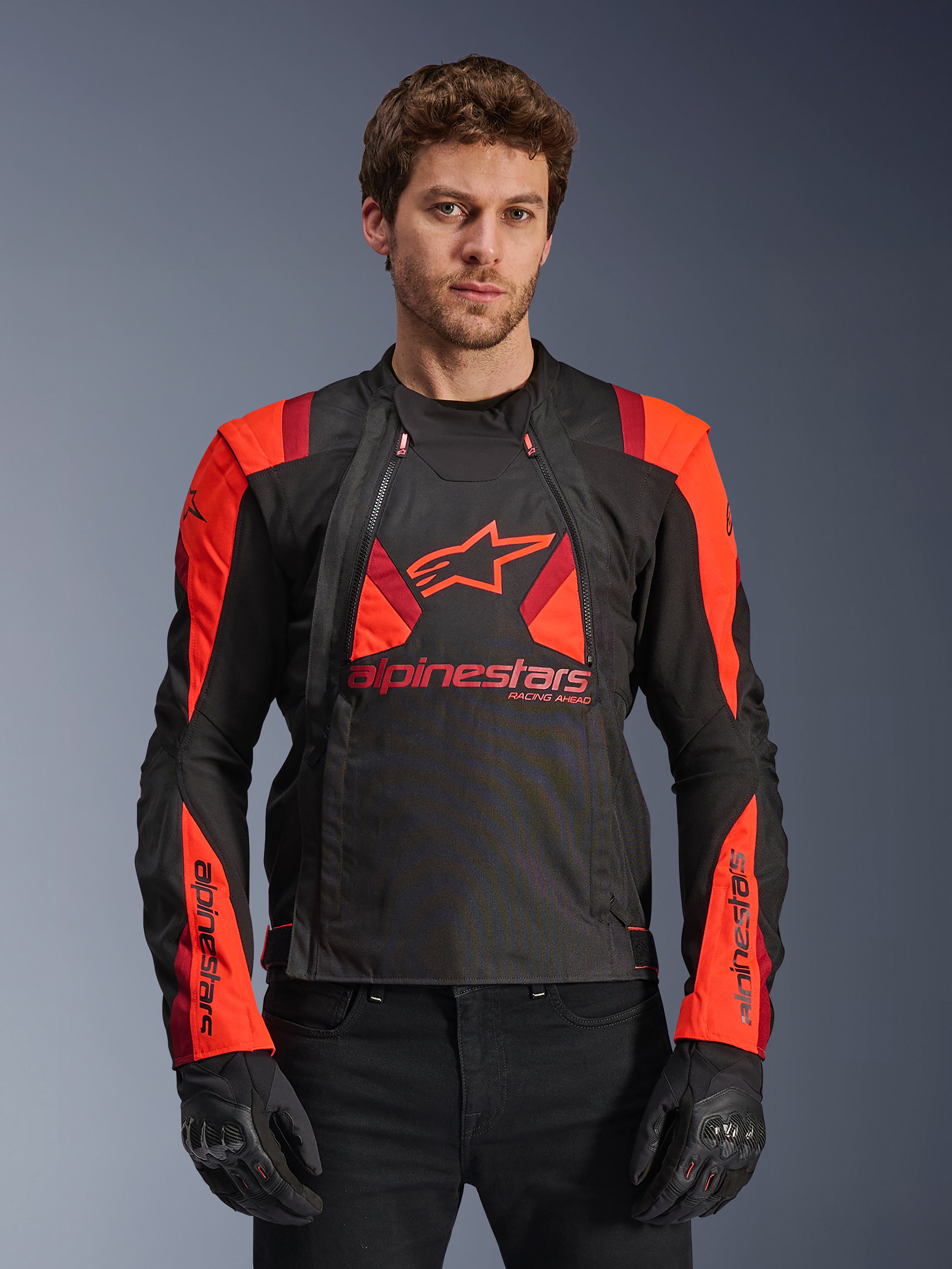 Alpinestars T-Stunt Air Jacket, tekstylna kurtka motocyklowa, czarna, jaskrawoczerwona i ciemnoczerwona, z dużym logo gwiazdy i panelami z siatki zapewniającymi przepływ powietrza, noszona przez modela w czarnych rękawicach