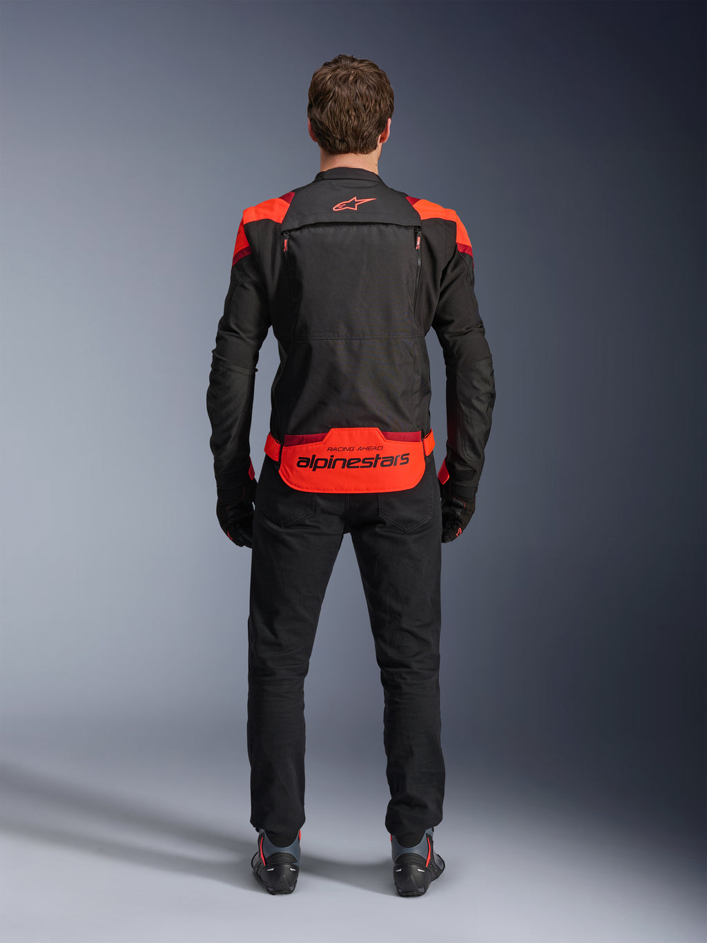 Kurtka Alpinestars T-Stunt Air, czarna z akcentami Red Fluo i ciemnoczerwonymi, widok z tyłu na modelu, w zestawieniu z czarnymi spodniami i butami motocyklowymi, wyraźne logo Alpinestars na dolnym panelu pleców, modułowa konstrukcja z siateczki zapewniająca wentylację