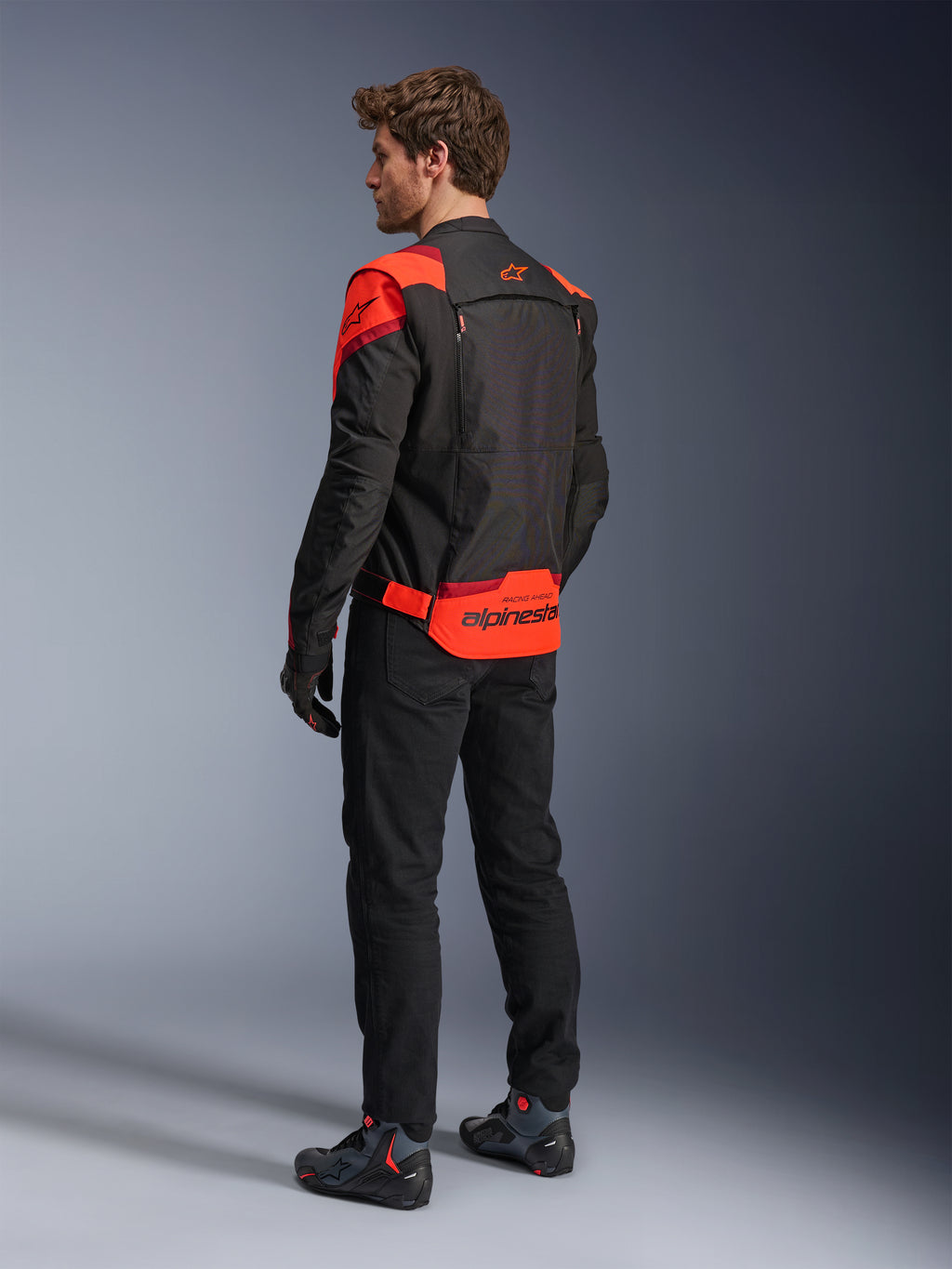 Mężczyzna w kurtce Alpinestars T-Stunt Air Jacket, widok z tyłu, kolor czarny, neonowy czerwony i ciemnoczerwony, tekstylna kurtka motocyklowa z panelami z siatki o sportowym kroju, z wyraźnym brandingiem Alpinestars na dole pleców, zestawiona z czarnymi spodniami i butami motocyklowymi