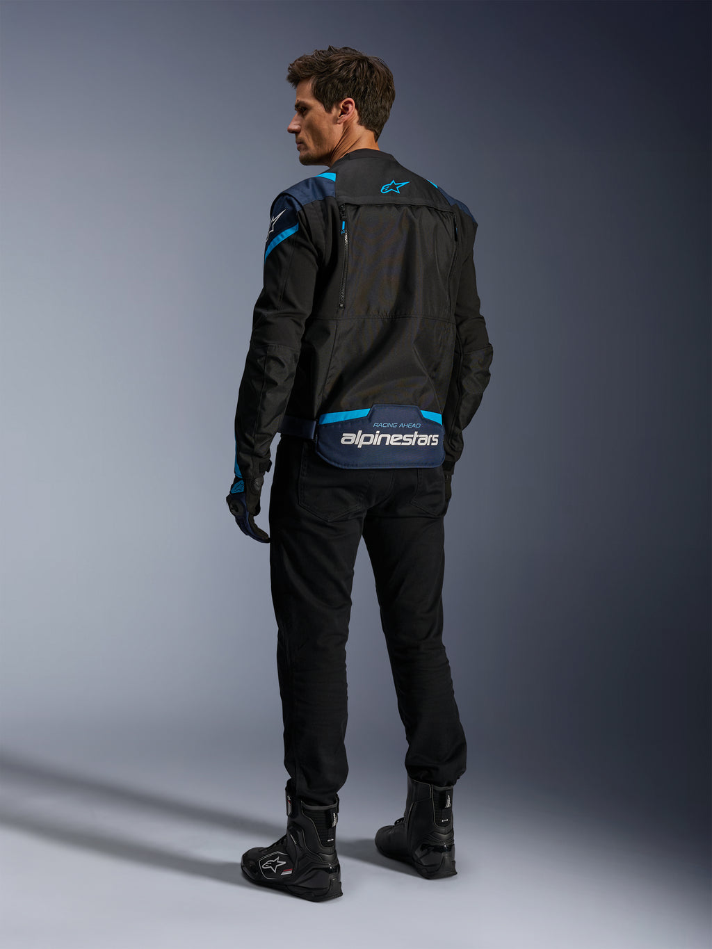 Kurtka Alpinestars T-Stunt Air, kurtka motocyklowa, czarna z ciemnoniebieskimi i błękitnymi akcentami, widok z tyłu pokazujący modułowe panele z siatki i logo na dolnej części pleców, noszona przez modela z czarnymi spodniami i butami motocyklowymi.