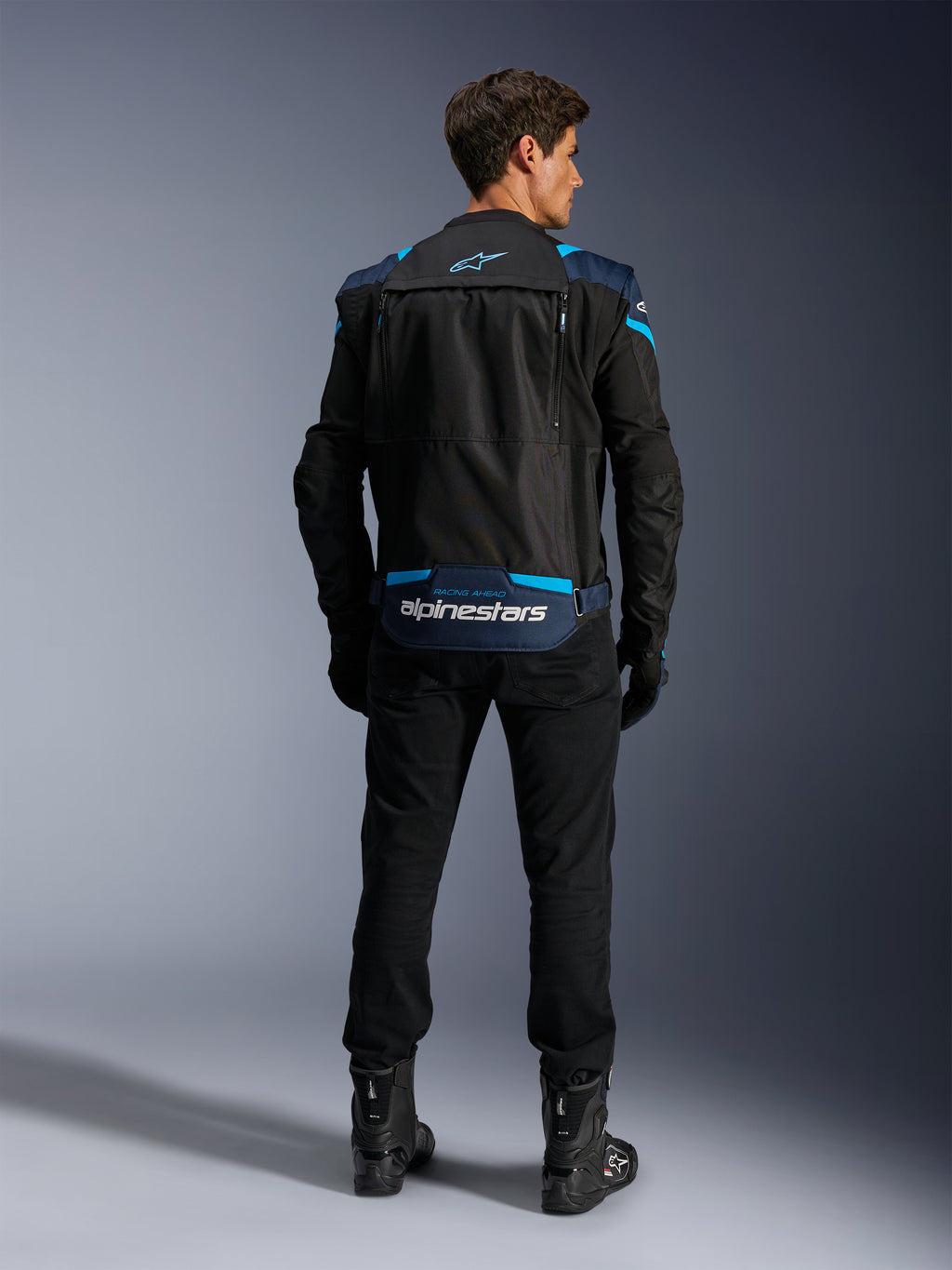 Alpinestars T-Stunt Air Jacket, modułowa tekstylna kurtka motocyklowa, czarna, ciemnoniebieska i jasnoniebieska, widok z tyłu pokazujący dolną kieszeń na plecach na rękawy z białym logo Alpinestars, noszona z czarnymi spodniami i butami.