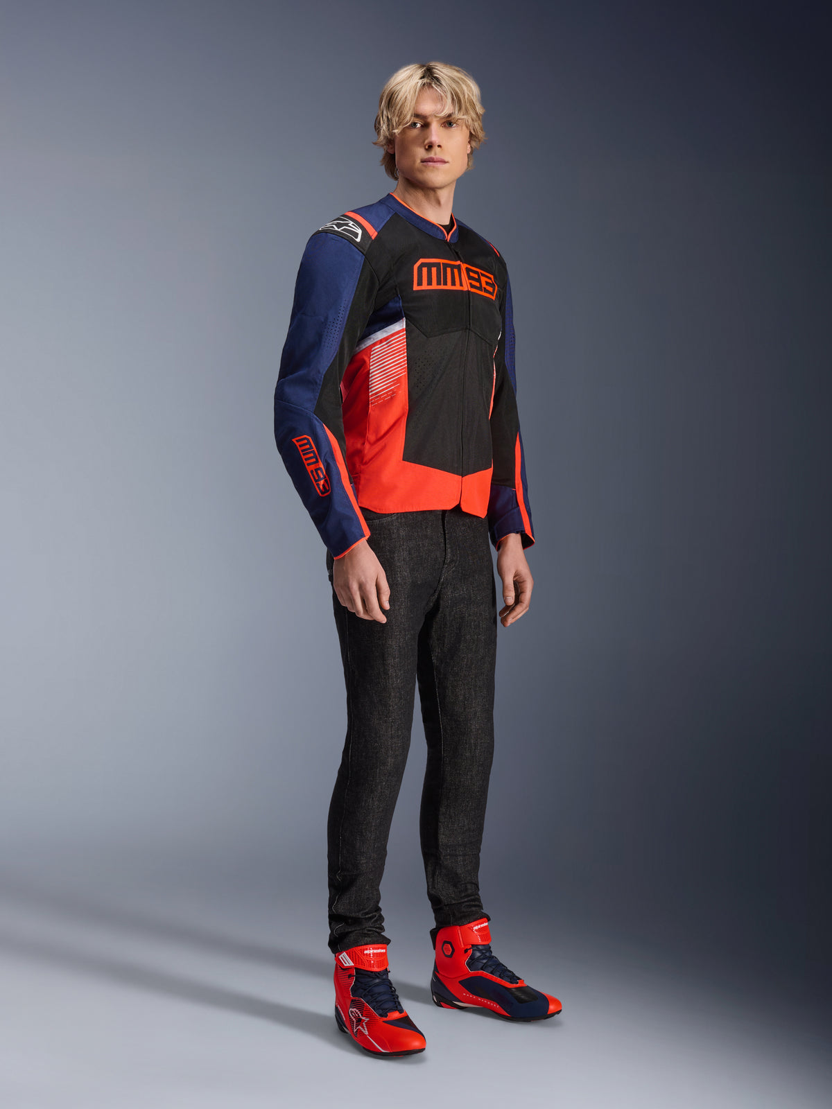 Męski model ubrany w kurtkę Alpinestars MM93 T-SPS Air V2 w kolorach granatowym, czarnym i czerwonym fluo, z wyrazistym brandingiem MM93 na klatce piersiowej i rękawach, w zestawieniu z ciemnymi dżinsami i pasującymi czerwono-niebieskimi butami motocyklowymi, sportowa miejska odzież motocyklowa