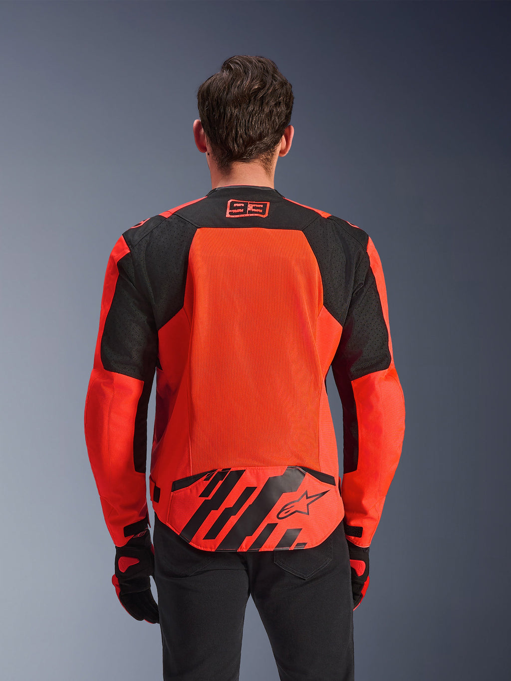 Kurtka Alpinestars MM93 T-SPS Air V2, widok z tyłu, kolor czerwony fluo i czarny, logo wyścigowe 93 na karku, duży panel z siatki na plecach dla optymalnego przepływu powietrza, dolna patka z logo gwiazdy Alpinestars i geometrycznymi pasami, noszona z czarnymi spodniami i rękawicami motocyklowymi