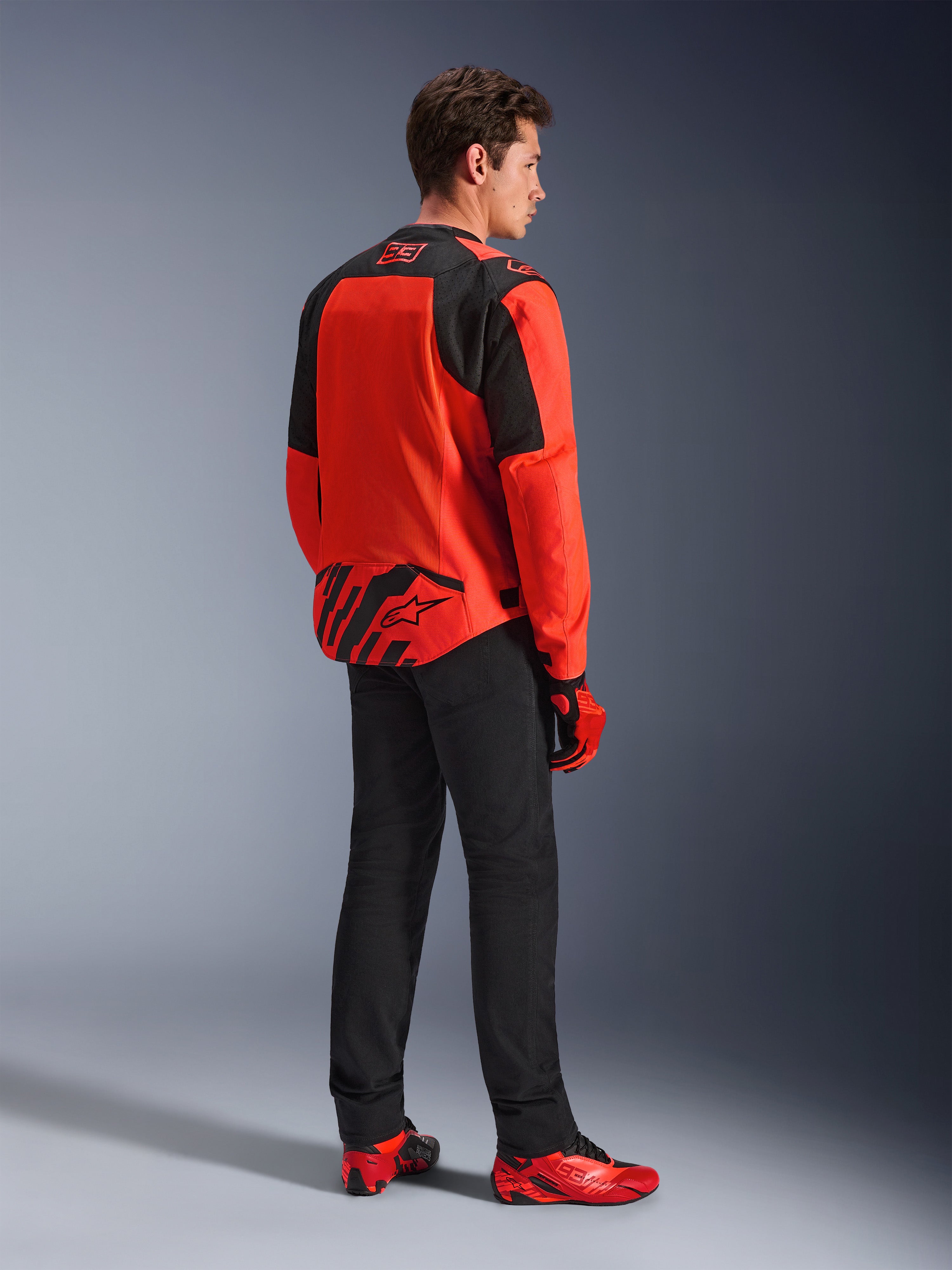 Kurtka Alpinestars MM93 T-SPS Air V2, kolor czerwony fluorescencyjny i czarny, widok z tyłu pod kątem trzech czwartych na modelu, branding Marc Marquez 93 na kołnierzu i w dolnej części pleców, czarne spodnie i czerwone buty motocyklowe.