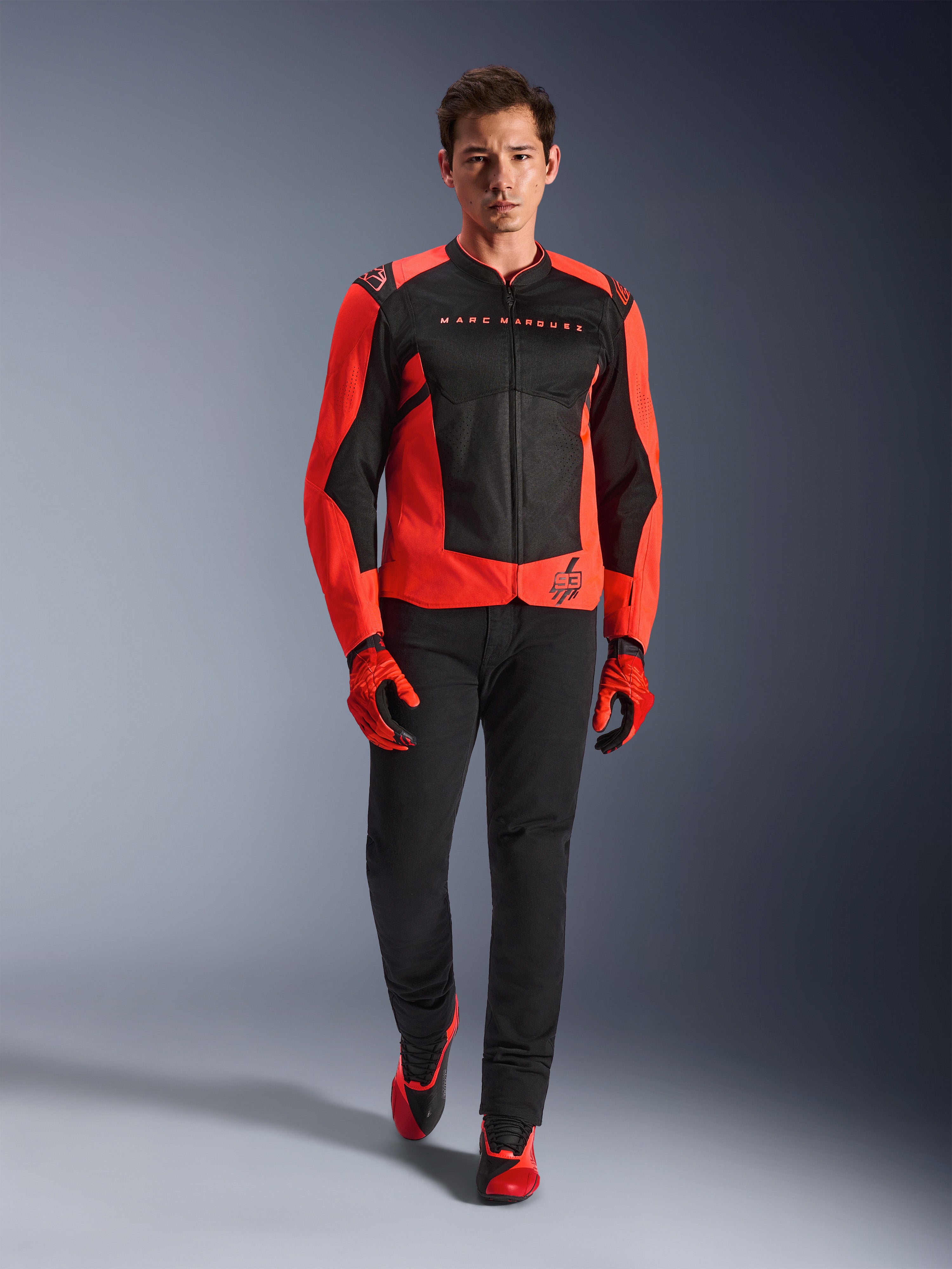 Alpinestars MM93 T-SPS Air V2 Jacket, czerwono-czarna (Red Fluo Black), noszona przez modela z dopasowanymi czerwonymi rękawicami i czarnymi spodniami, z brandingiem Marca Marqueza i logo 93, wentylowana sportowa kurtka motocyklowa do jazdy miejskiej