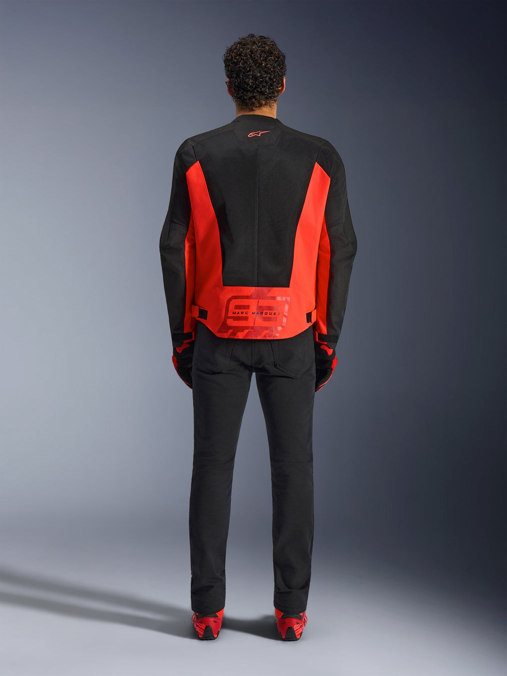 Alpinestars MM93 C-1 Air Jacket, kurtka motocyklowa, jaskrawoczerwona (fluo) i czarna, widok z tyłu z grafiką Marc Marquez 93 na dole pleców, techniczne panele siatkowe dla wentylacji, w zestawieniu z czarnymi spodniami i akcesoriami z czerwonymi akcentami