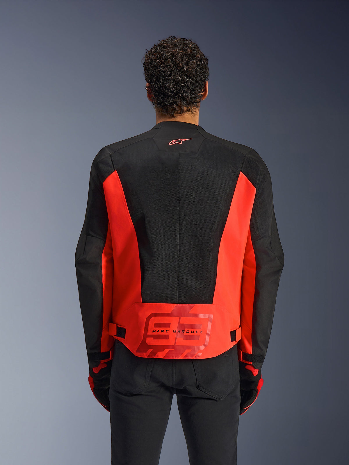 Widok z tyłu mężczyzny w kurtce Alpinestars MM93 C-1 Air Jacket w kolorze Red Fluo i czarnym, z dużym logo Marca Marqueza „93” na dole pleców, lekkimi panelami z siatki dla wentylacji, w zestawie z czarnymi spodniami i rękawicami.