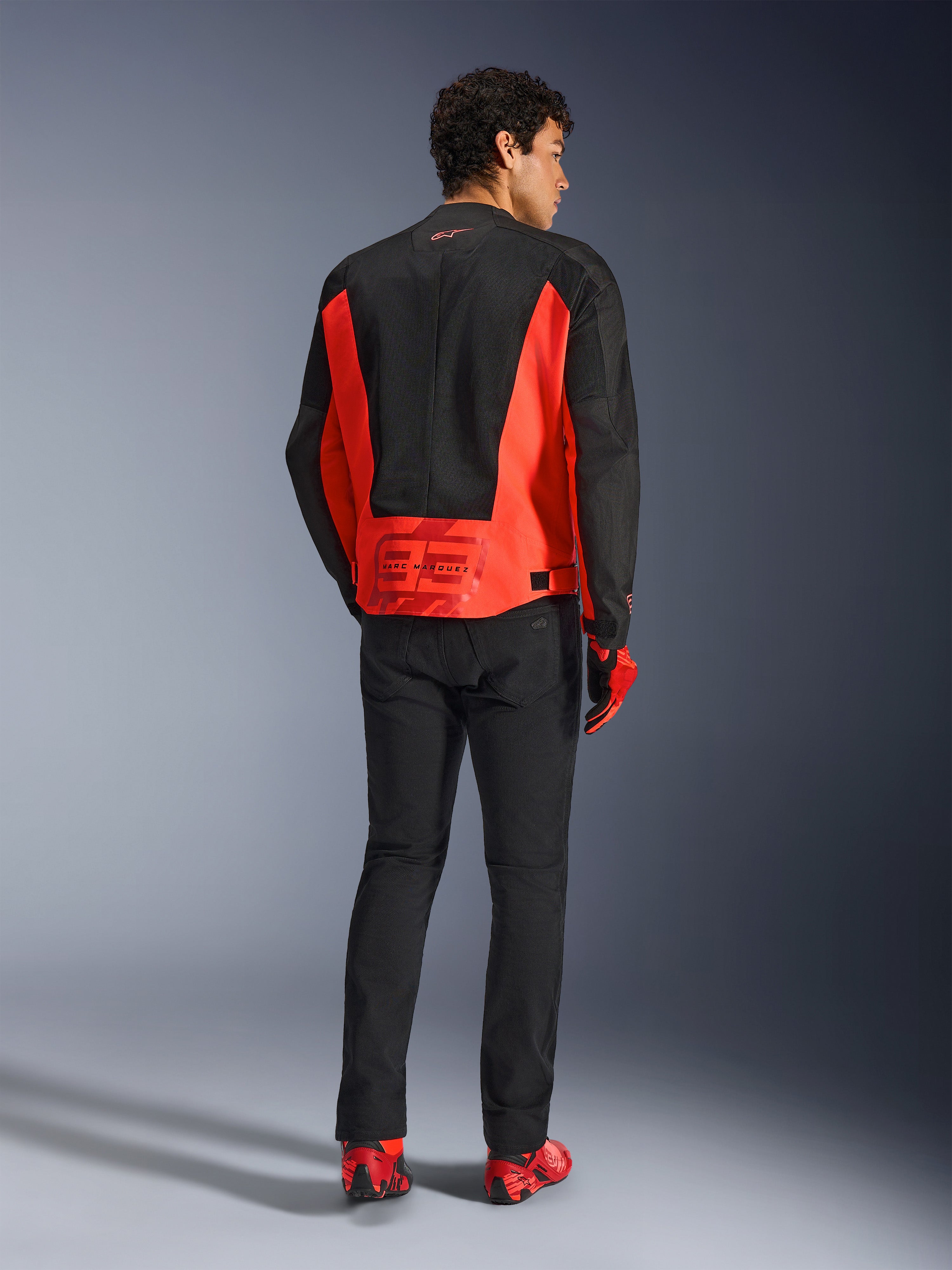 Alpinestars MM93 C-1 Air Jacket, widok z tyłu modela w kurtce w kolorze czerwonym fluo i czarnym, logo Marc Marquez 93 na dole pleców, czarne spodnie i czerwone buty motocyklowe, miejski styl motocyklowy