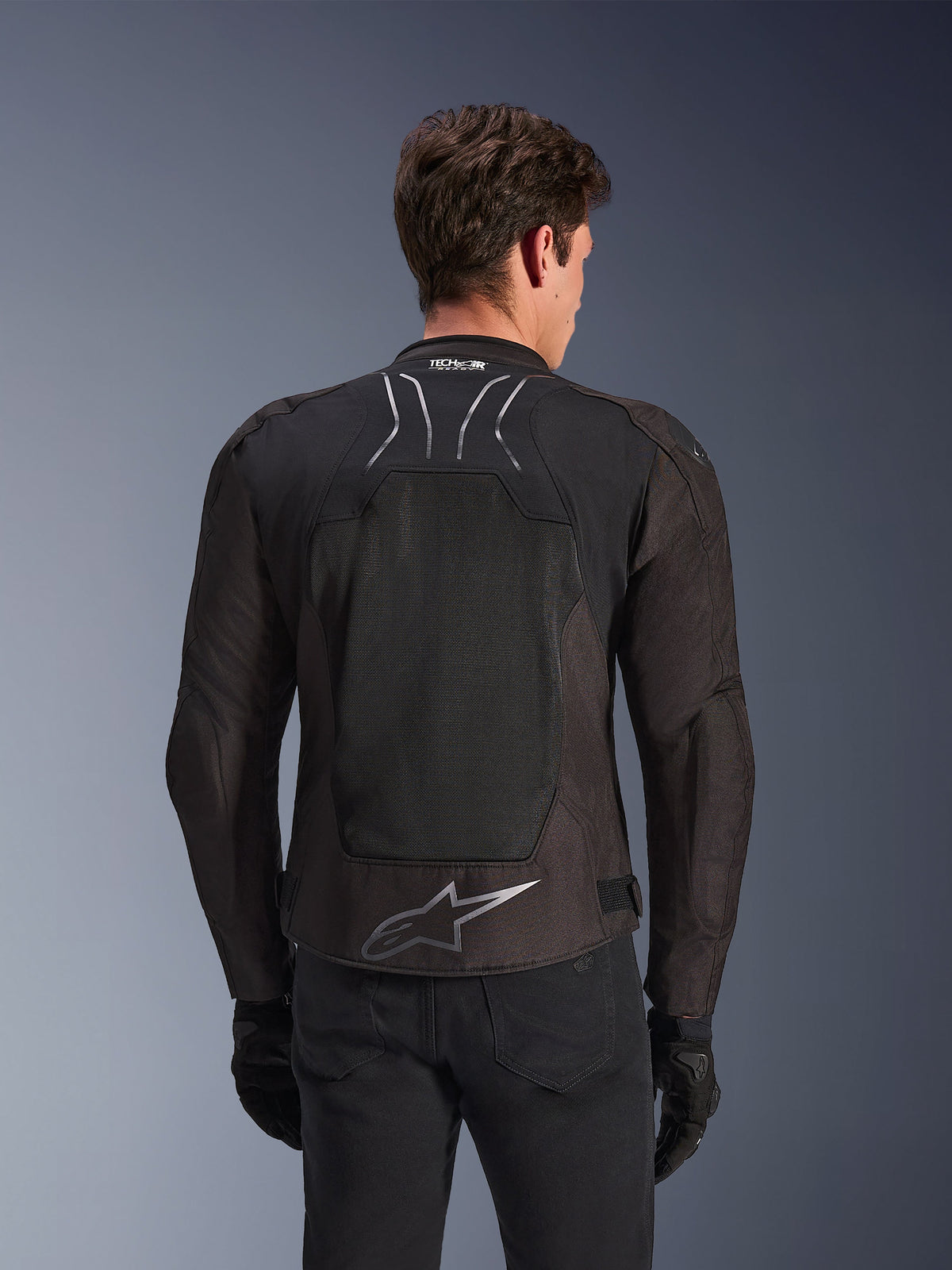 Kurtka Alpinestars SMX Air w kolorze Black Anthracite, widok z tyłu na modelu, duży panel z siatki technicznej na plecach zapewniający wentylację, oznaczenie Tech-Air Ready na kołnierzu, srebrne logo Astar na dole pleców, czarne rękawice i spodnie motocyklowe