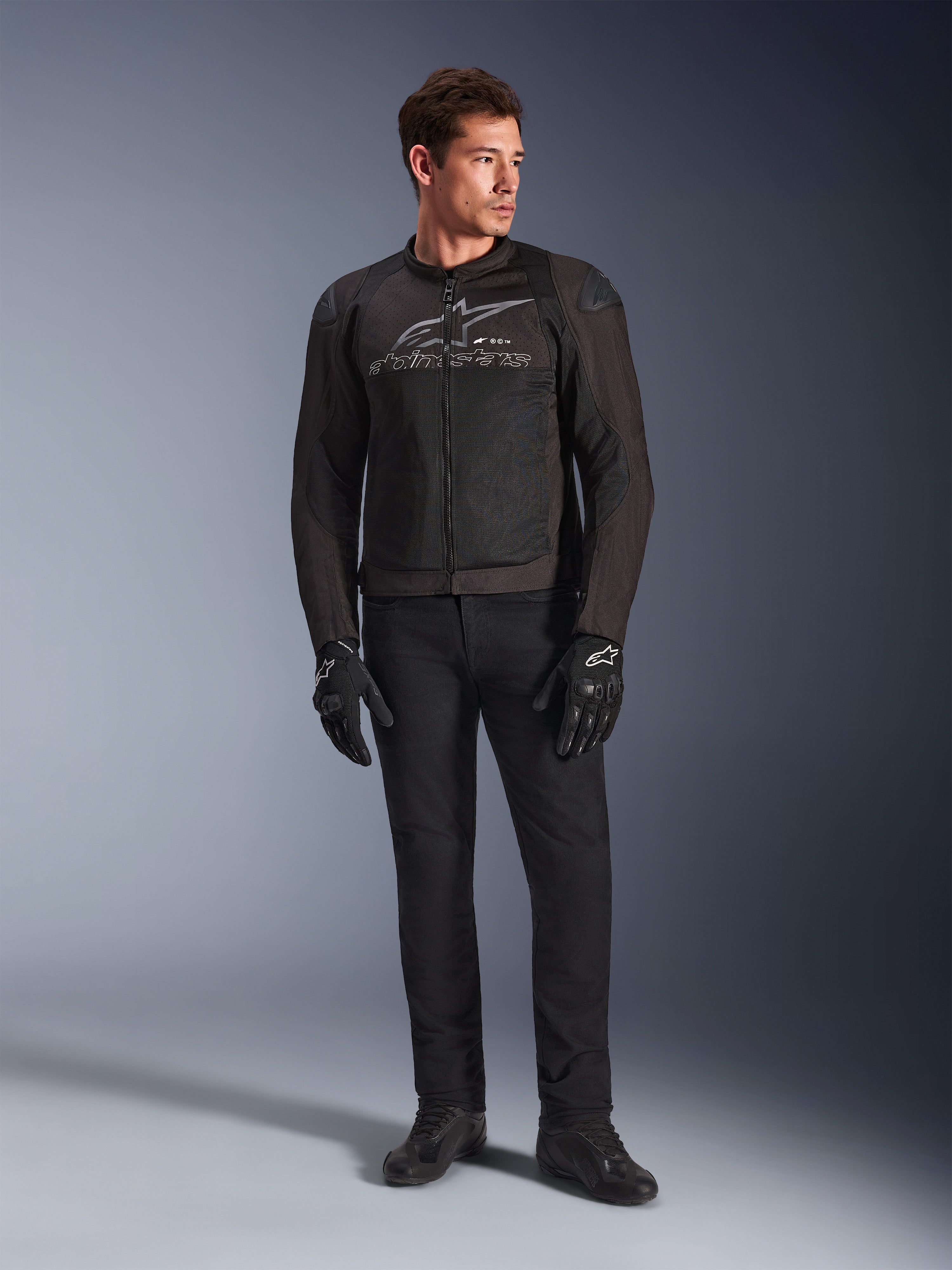 Męski model w kurtce motocyklowej Alpinestars SMX Air, kolor Black Anthracite, wentylowane panele z siatki i szare logo na klatce piersiowej, czarne spodnie i rękawice ochronne, tło studyjne z gradientem