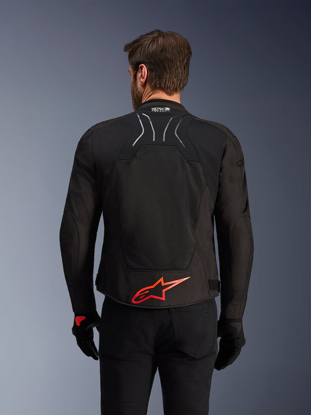 Kurtka Alpinestars SMX Air, widok z tyłu, czarna z jaskrawoczerwonym logo Alpinestars na dole pleców, z technicznym panelem z siatki o wysokim przepływie powietrza i oznaczeniem Tech-Air Ready, noszona przez motocyklistę w czarnych spodniach i rękawicach