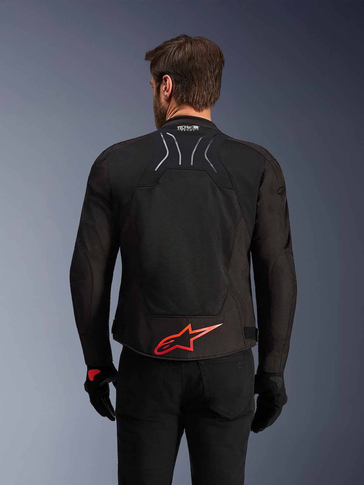 Kurtka Alpinestars SMX Air, widok z tyłu, czarna z jaskrawoczerwonym logo Alpinestars na dole pleców, z technicznym panelem z siatki o wysokim przepływie powietrza i oznaczeniem Tech-Air Ready, noszona przez motocyklistę w czarnych spodniach i rękawicach