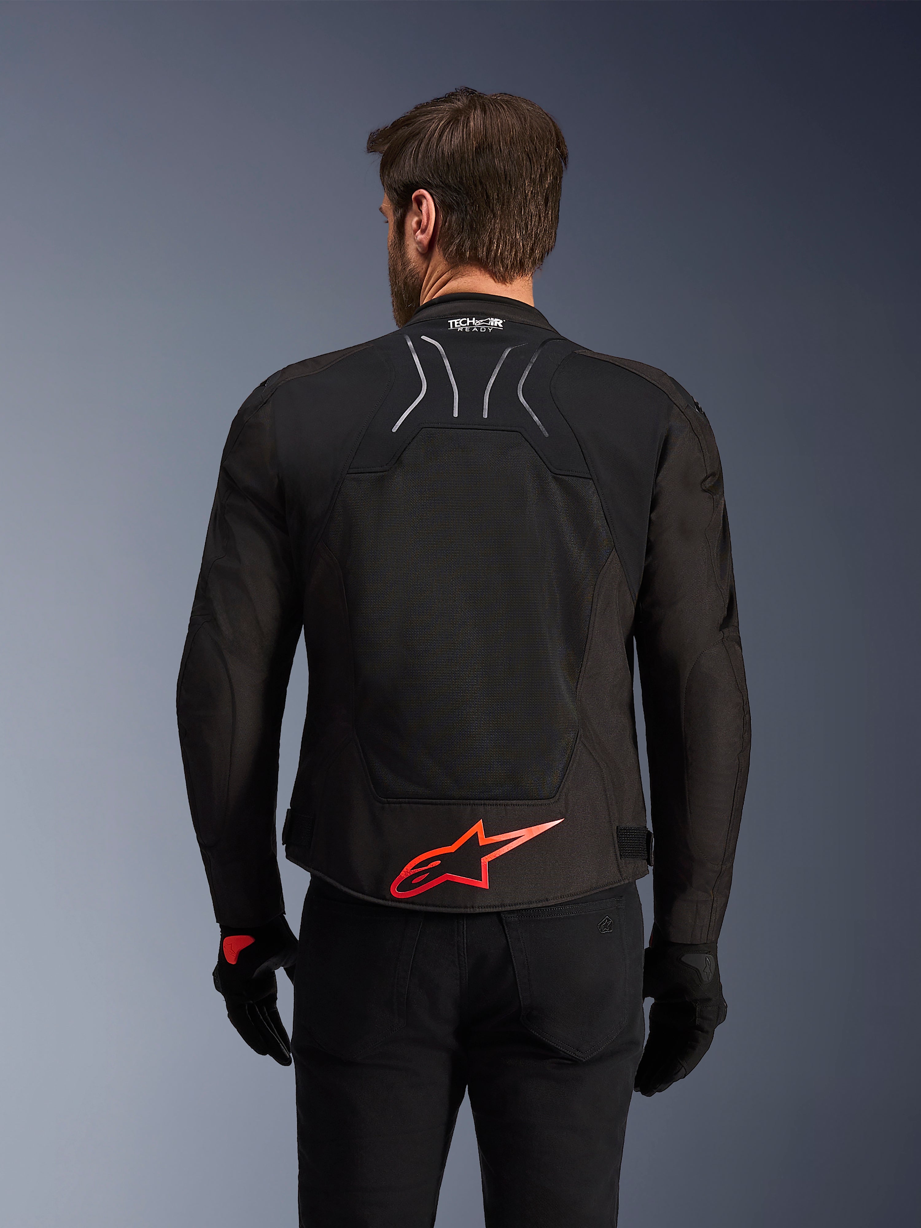 Kurtka Alpinestars SMX Air, widok z tyłu, czarna z jaskrawoczerwonym logo Alpinestars na dole pleców, z technicznym panelem z siatki o wysokim przepływie powietrza i oznaczeniem Tech-Air Ready, noszona przez motocyklistę w czarnych spodniach i rękawicach