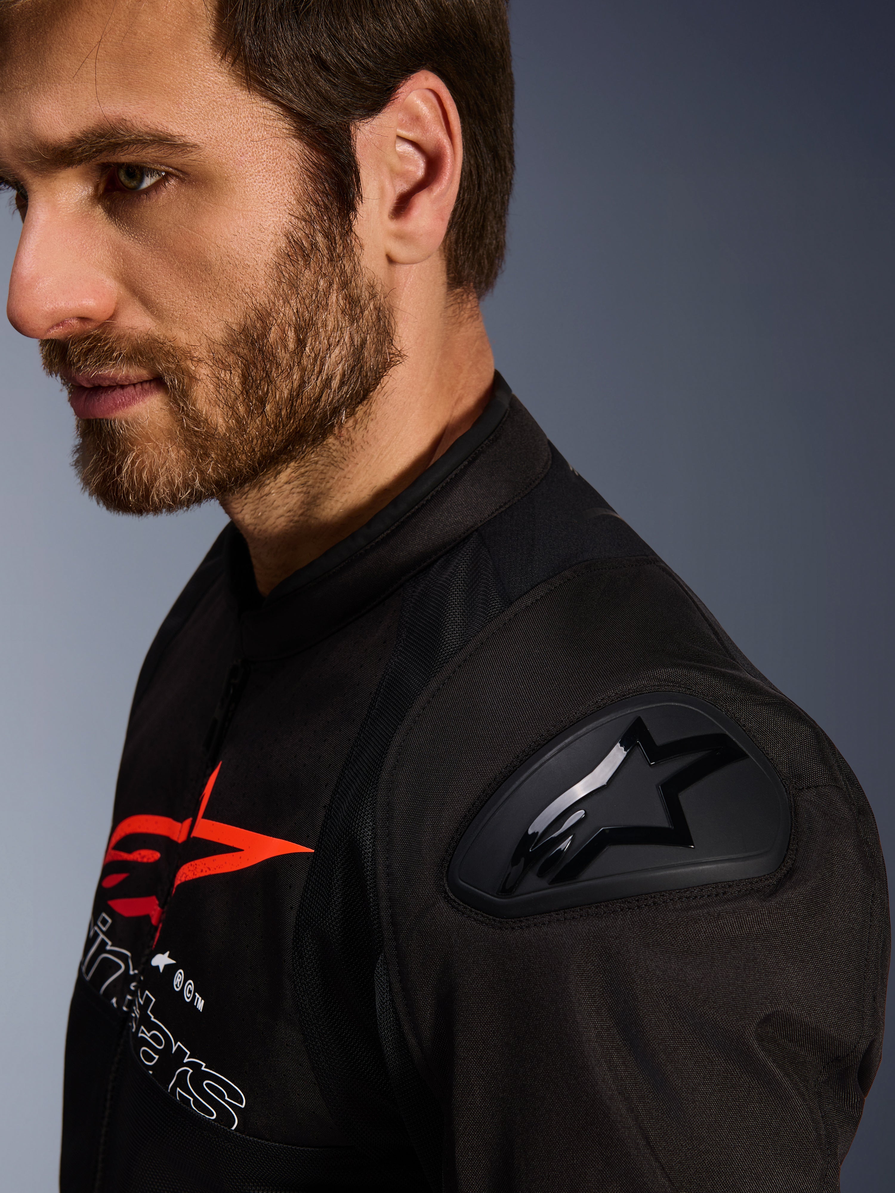 Alpinestars SMX Air Jacket, czarno-czerwona, zbliżenie na ochraniacz ramion DFS Lite z logo gwiazdy, noszona przez mężczyznę z brodą, tekstylna sportowa i wyścigowa kurtka motocyklowa.
