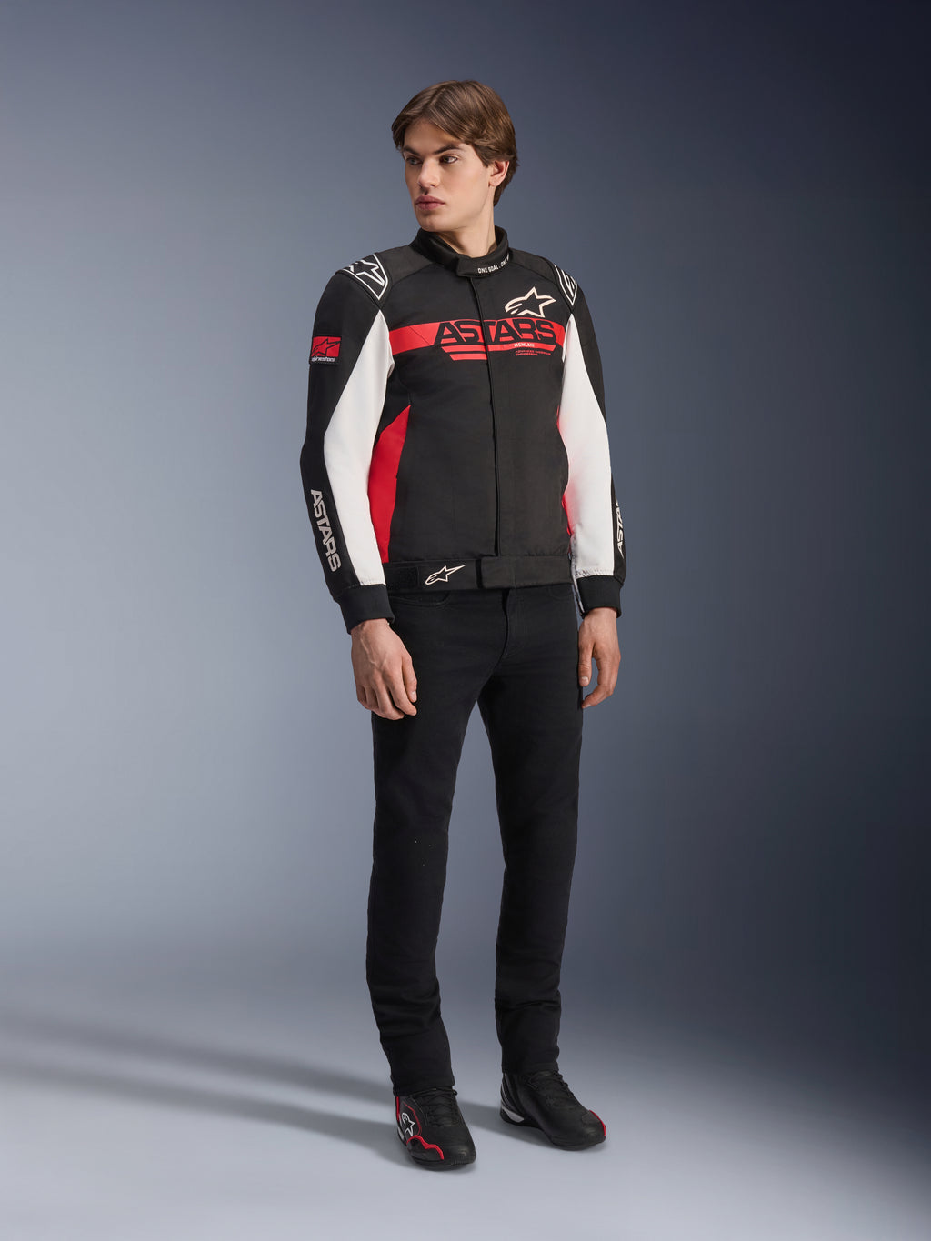 Alpinestars Monza Sport Jacket, czarno-czerwono-biała tekstylna kurtka motocyklowa, design inspirowany wyścigami z brandingiem ASTARS na klatce piersiowej i rękawach, noszona przez modela z czarnymi spodniami i butami motocyklowymi, casualowa odzież motocyklowa w sportowym stylu