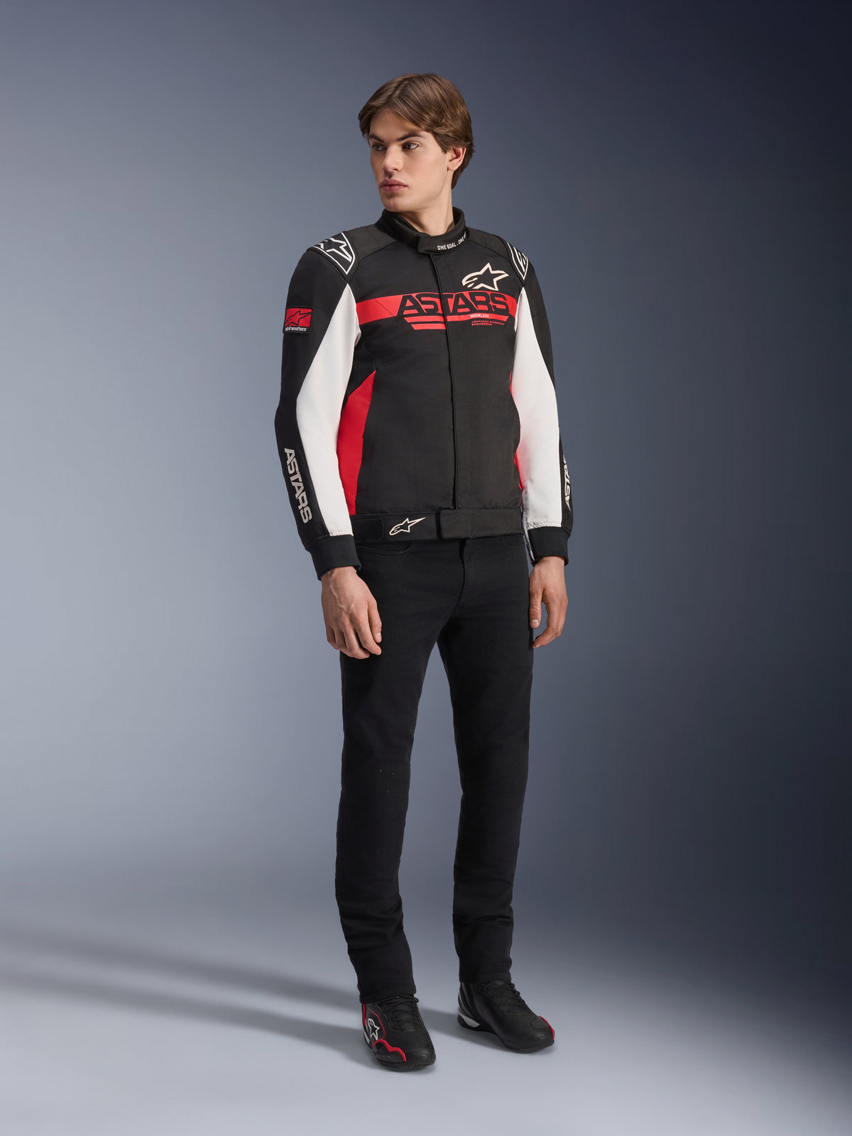 Alpinestars Monza Sport Jacket, czarno-czerwono-biała tekstylna kurtka motocyklowa, design inspirowany wyścigami z brandingiem ASTARS na klatce piersiowej i rękawach, noszona przez modela z czarnymi spodniami i butami motocyklowymi, casualowa odzież motocyklowa w sportowym stylu