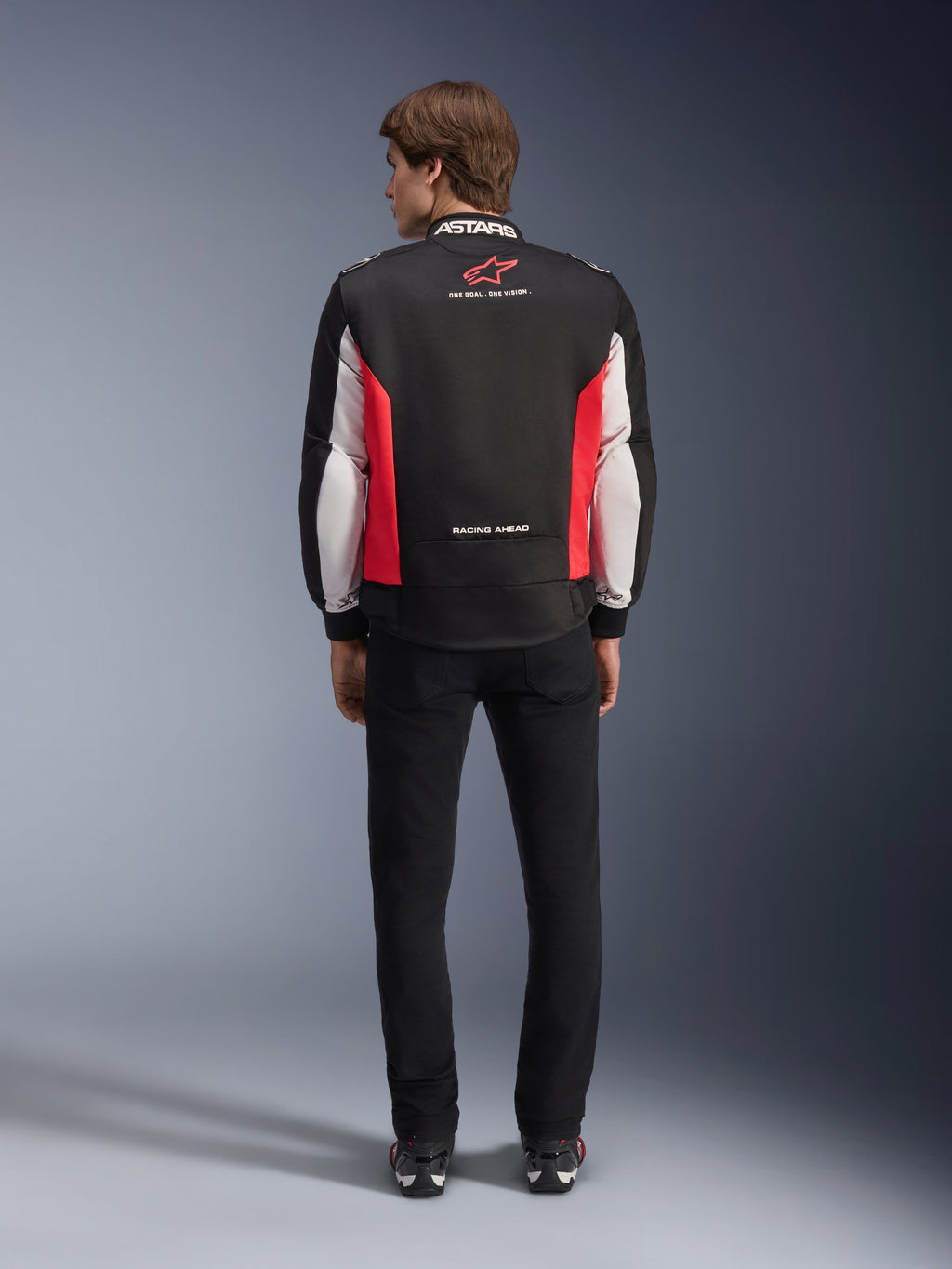 Kurtka Alpinestars Monza Sport, czarno-czerwono-biała tekstylna kurtka motocyklowa, widok z tyłu na modelu, czerwone logo gwiazdy i napis „ASTARS” na kołnierzu z hasłami, zestawiona z czarnymi spodniami i butami motocyklowymi.
