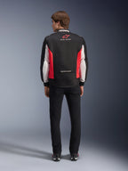 Kurtka Alpinestars Monza Sport, czarno-czerwono-biała tekstylna kurtka motocyklowa, widok z tyłu na modelu, czerwone logo gwiazdy i napis „ASTARS” na kołnierzu z hasłami, zestawiona z czarnymi spodniami i butami motocyklowymi.