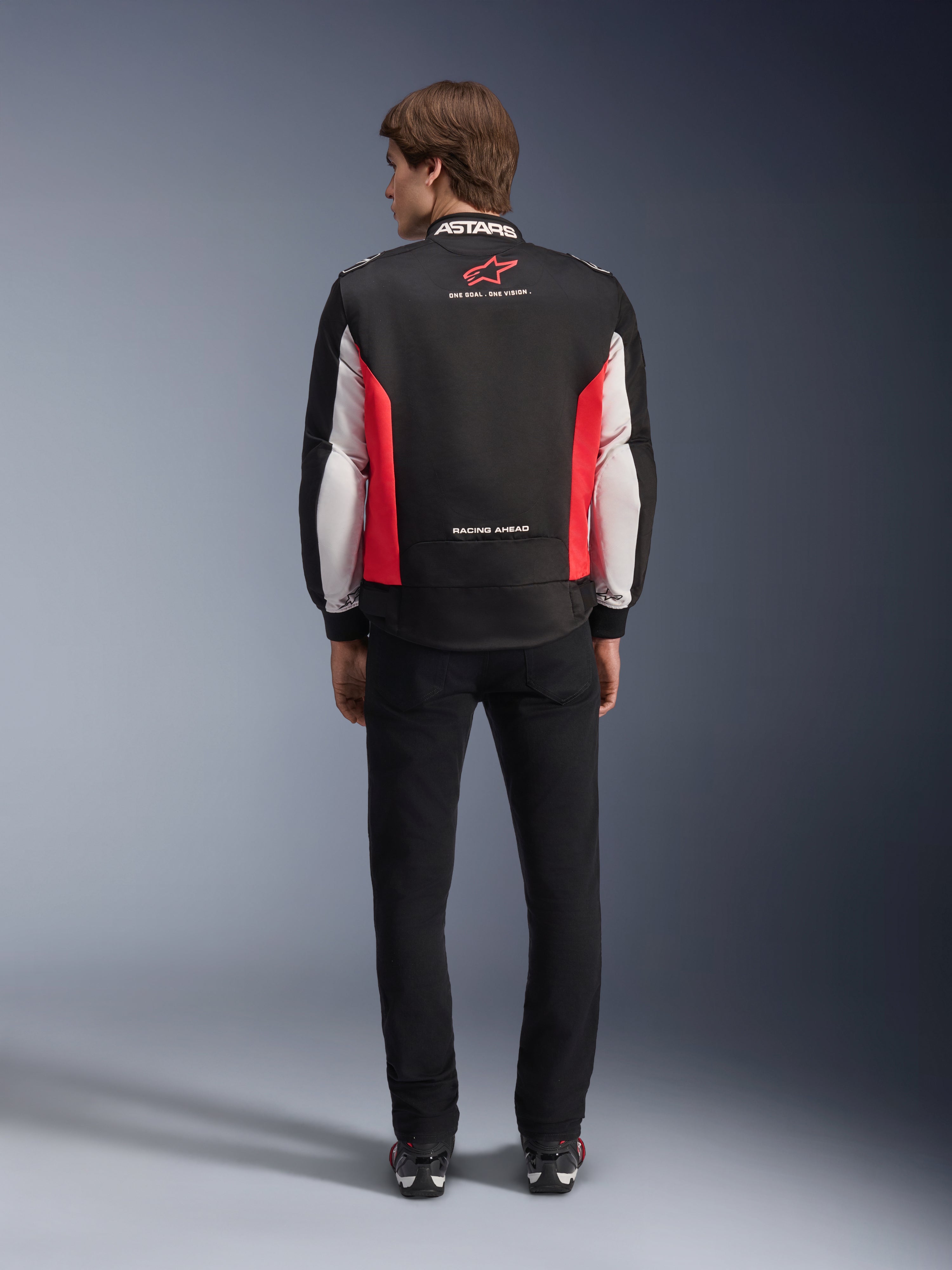 Kurtka Alpinestars Monza Sport, czarno-czerwono-biała tekstylna kurtka motocyklowa, widok z tyłu na modelu, czerwone logo gwiazdy i napis „ASTARS” na kołnierzu z hasłami, zestawiona z czarnymi spodniami i butami motocyklowymi.