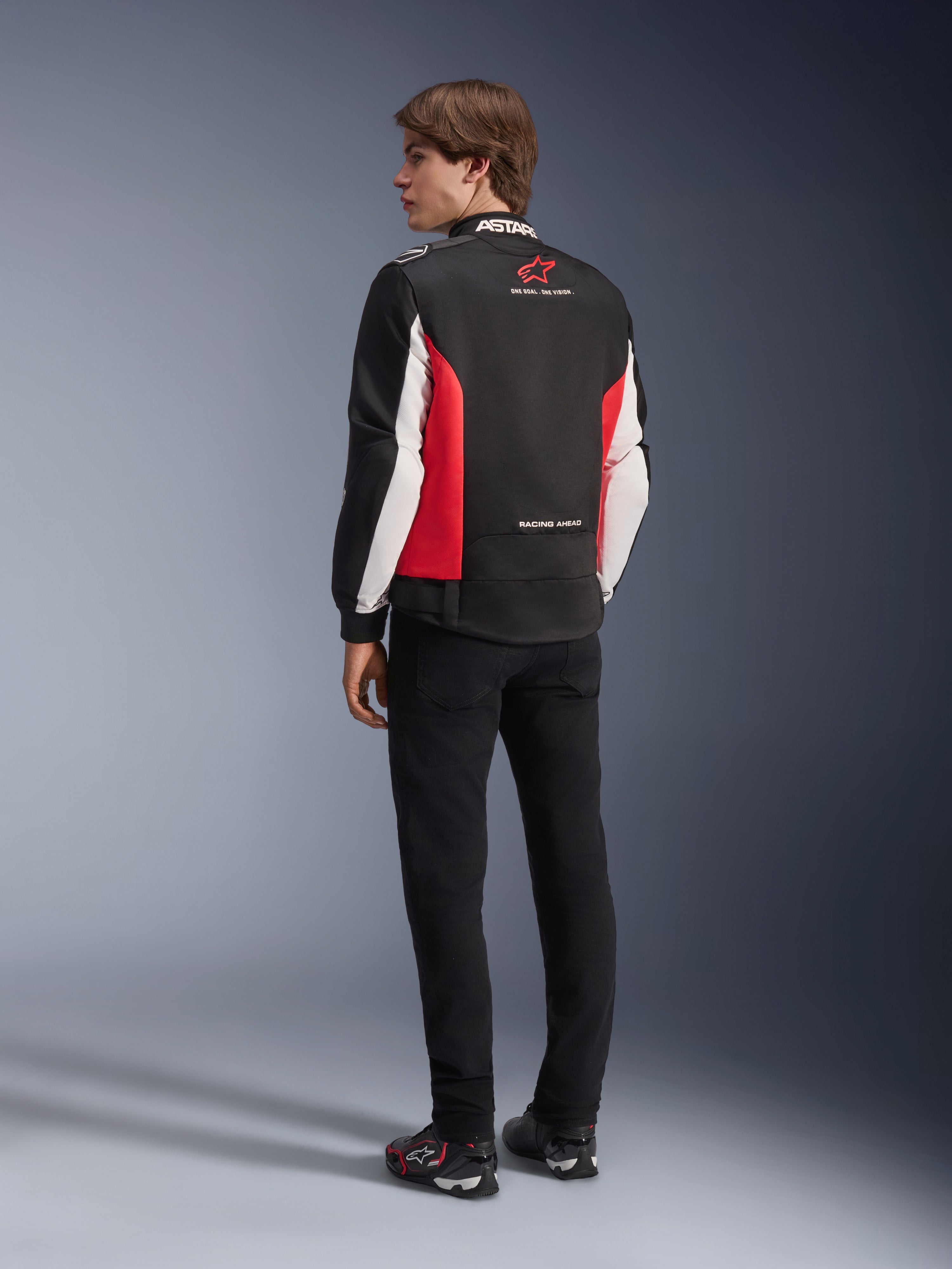 Kurtka Alpinestars Monza Sport, tekstylna kurtka motocyklowa, czarna z jaskrawoczerwonymi i białymi elementami, widok modelu od tyłu z czerwonymi panelami bocznymi i białymi akcentami na rękawach, logo Alpinestars i napis „ONE GOAL. ONE VISION.”, zestawiona z czarnymi spodniami i butami motocyklowymi w sportowym stylu