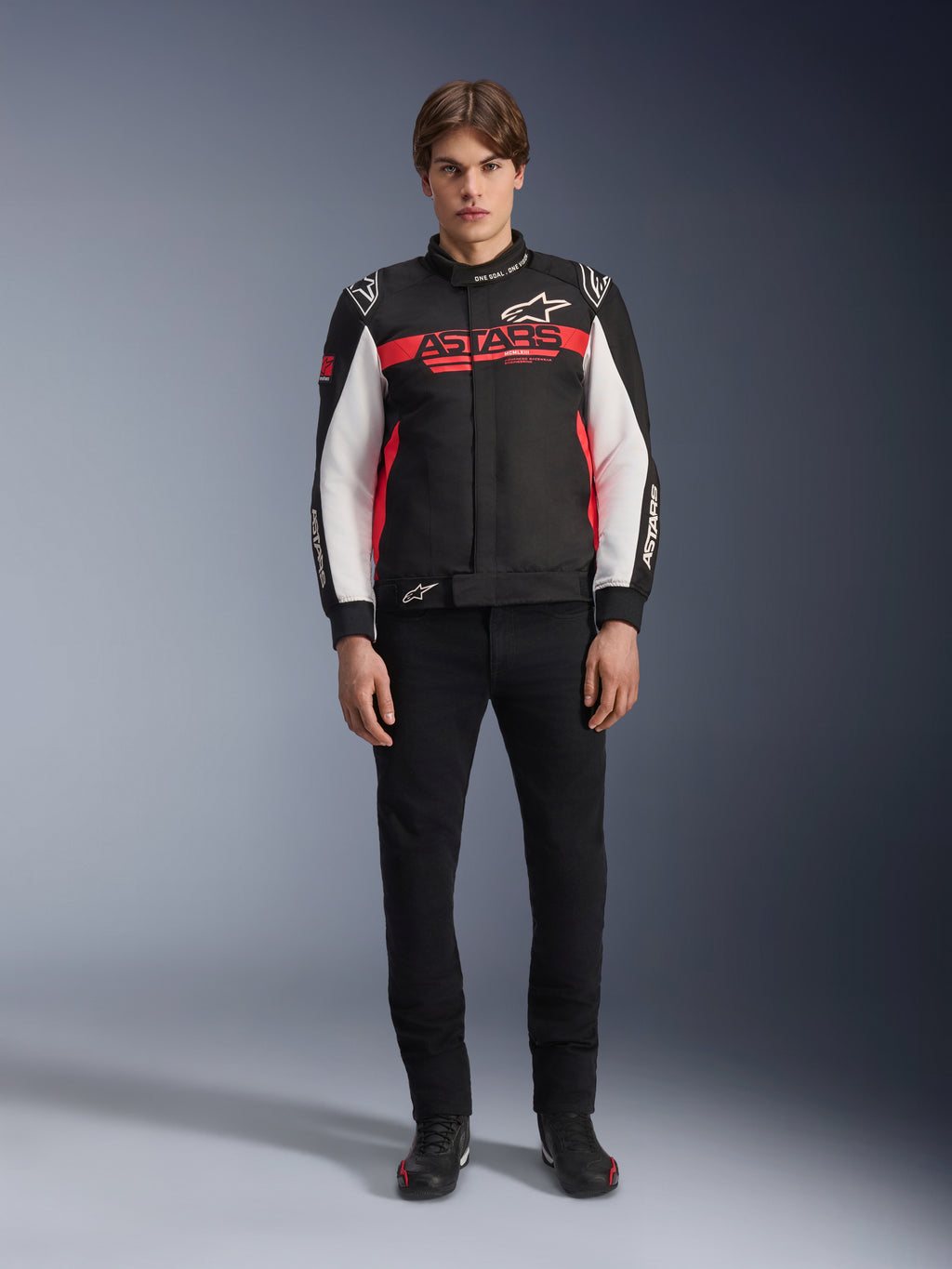 Męski model w kurtce Alpinestars Monza Sport w kolorze czarnym, jaskrawoczerwonym i białym, z dużym czerwonym logo „ASTARS” na klatce piersiowej i białymi panelami na rękawach, w zestawieniu z czarnymi spodniami i czarnymi butami motocyklowymi.