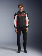 Męski model w kurtce Alpinestars Monza Sport w kolorze czarnym, jaskrawoczerwonym i białym, z dużym czerwonym logo „ASTARS” na klatce piersiowej i białymi panelami na rękawach, w zestawieniu z czarnymi spodniami i czarnymi butami motocyklowymi.