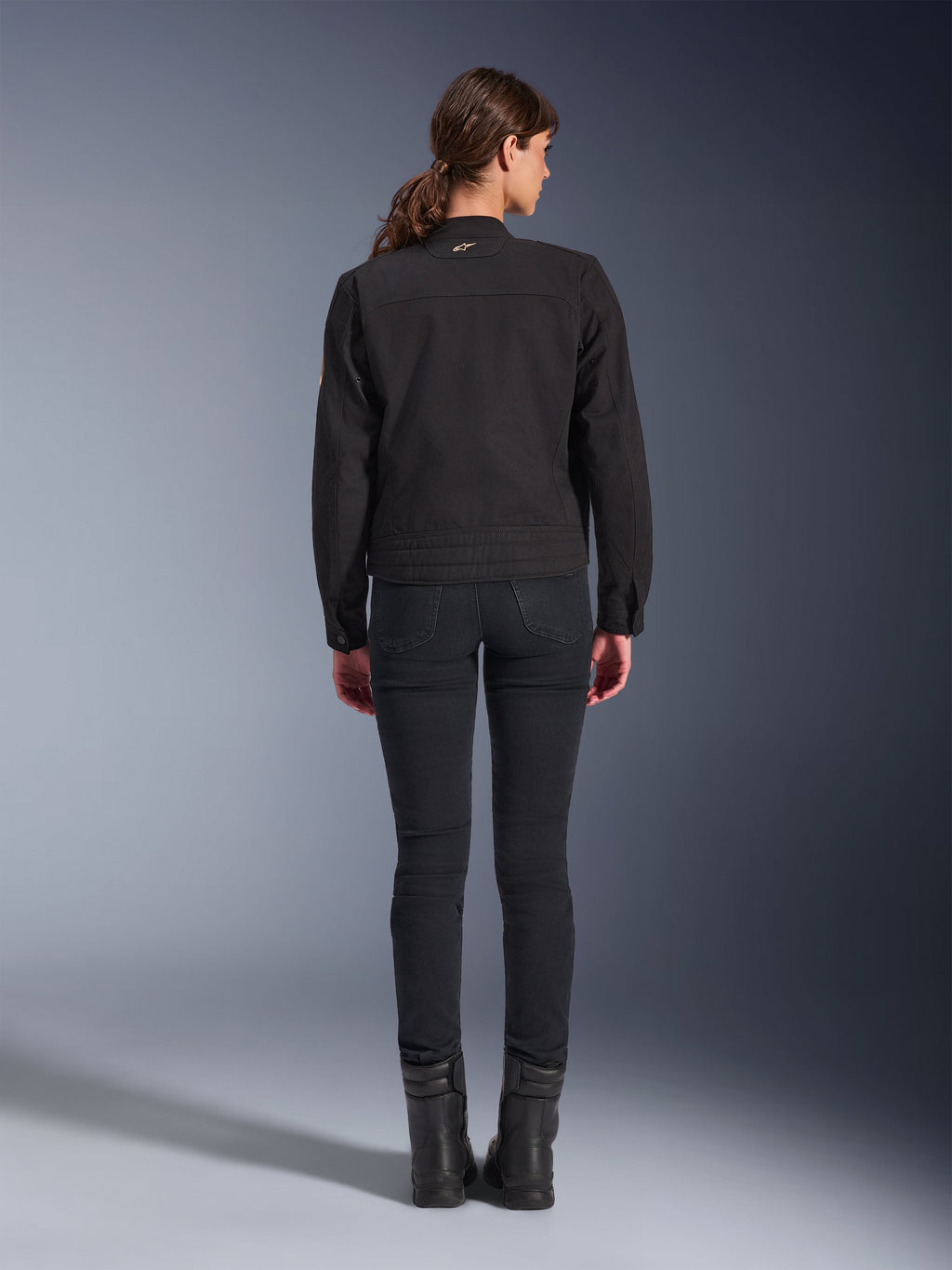 Kurtka Alpinestars Stella Grace, damska miejska kurtka motocyklowa, czarna, widok z tyłu prezentujący design inspirowany odzieżą roboczą, noszona z czarnymi dopasowanymi spodniami i czarnymi butami, zdjęcie studyjne