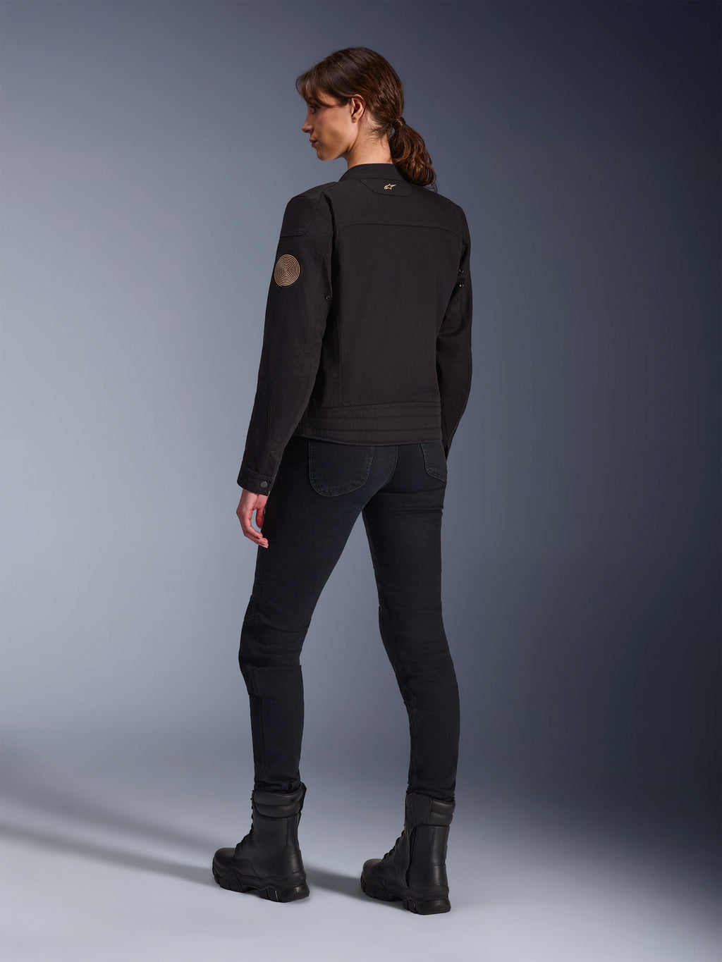 Modelka w czarnej kurtce Alpinestars Stella Grace, widok z tyłu uwydatniający spiralną naszywkę Oscar w stylu vintage na ramieniu oraz małe logo z gwiazdą na kołnierzu, stylizacja z czarnymi spodniami motocyklowymi i czarnymi butami do jazdy miejskiej