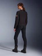 Modelka w czarnej kurtce Alpinestars Stella Grace, widok z tyłu uwydatniający spiralną naszywkę Oscar w stylu vintage na ramieniu oraz małe logo z gwiazdą na kołnierzu, stylizacja z czarnymi spodniami motocyklowymi i czarnymi butami do jazdy miejskiej