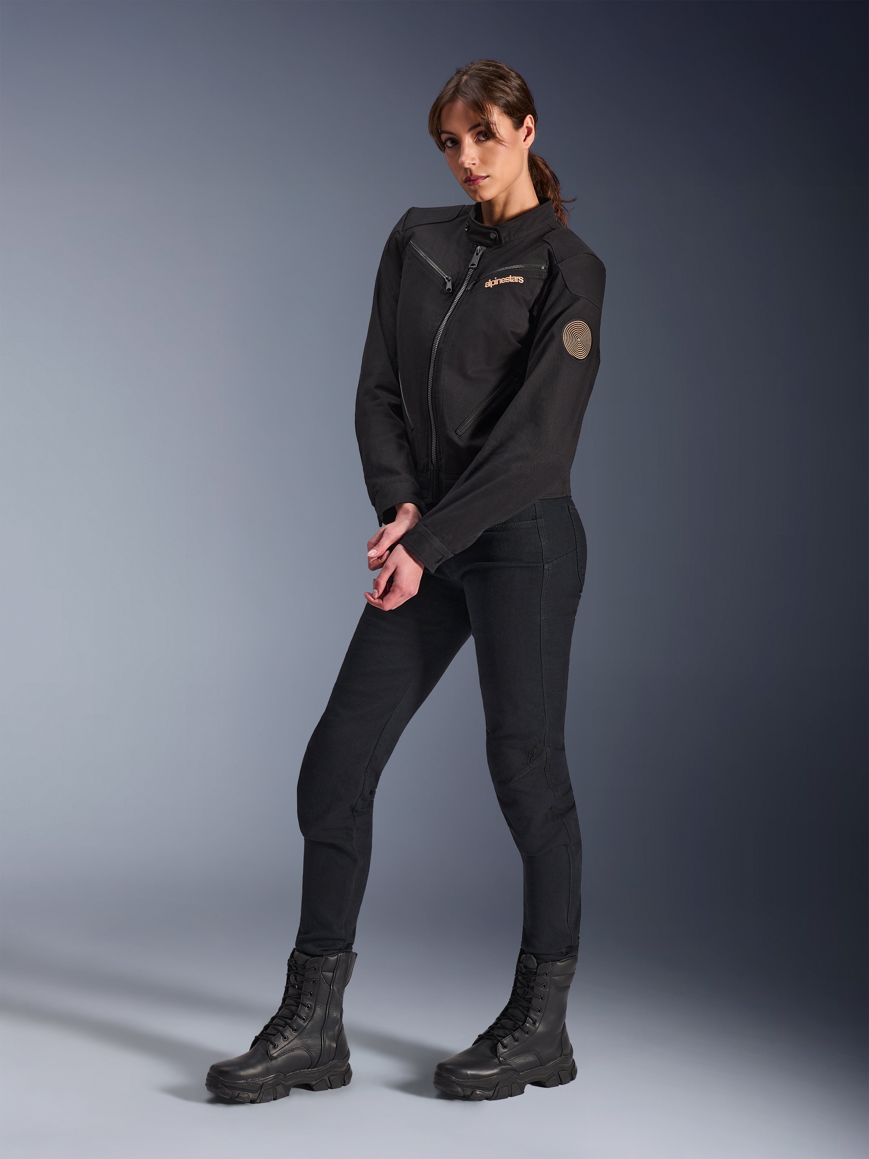 Modelka w kurtce Alpinestars Stella Grace, czarna miejska kurtka motocyklowa, w stylizacji z czarnymi spodniami i butami motocyklowymi, design inspirowany odzieżą roboczą, brązowe haftowane logo na piersi i okrągła spiralna naszywka na rękawie
