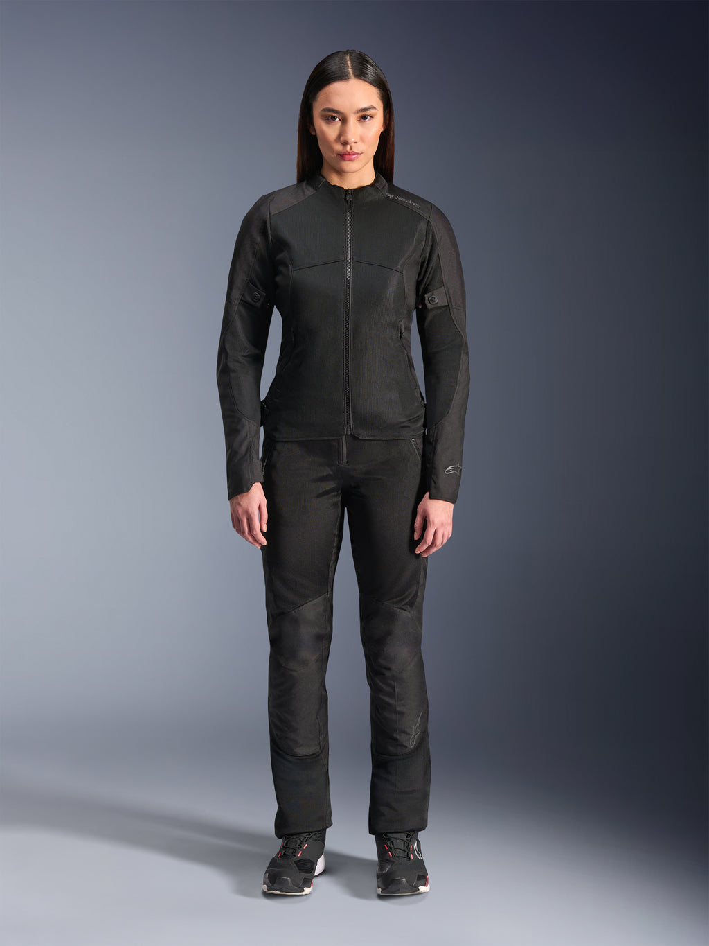 Kobieta ubrana w kurtkę Alpinestars Stella C-1 Air w kolorze czarnym, zestawioną z dopasowanymi czarnymi spodniami tekstylnymi i butami motocyklowymi, o smukłym miejskim designie z dużymi panelami z siatki zapewniającymi przepływ powietrza.