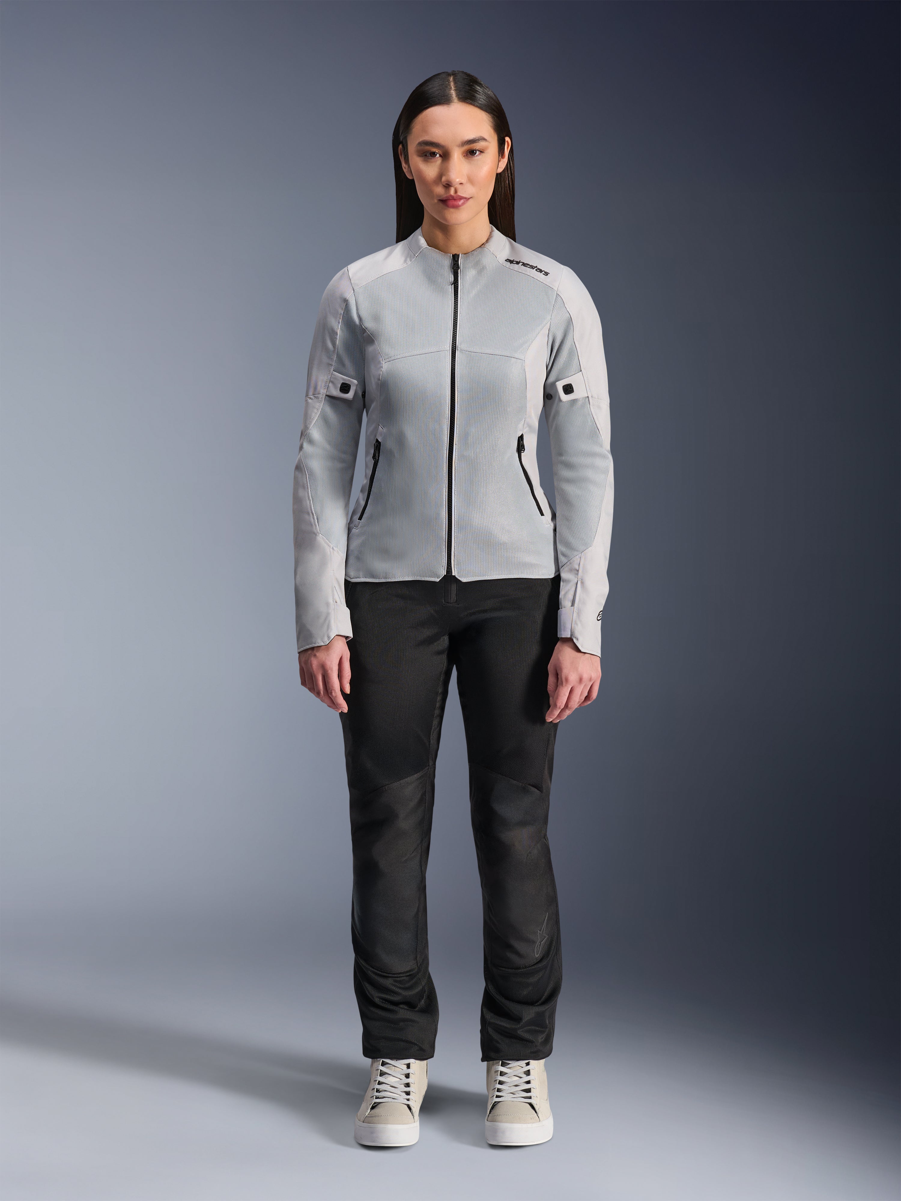 Kobieta w kurtce Alpinestars Stella C-1 Air w kolorze srebrnym, widok z przodu, miejska motocyklowa kurtka tekstylna z panelami z siatki technicznej, zestawiona z czarnymi spodniami i białymi trampkami, nowoczesna odzież motocyklowa