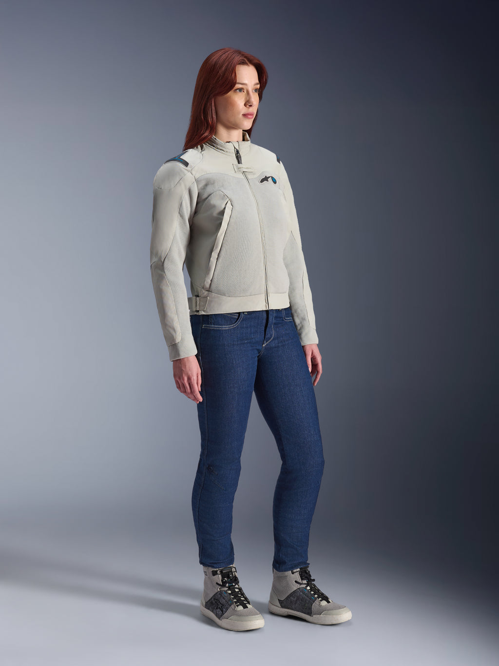 Kobieta ubrana w kurtkę Alpinestars Stella Circal-1 Air w kolorze Metal Gray i Methyl Blue, zestawioną z niebieskimi jeansami i codziennymi butami motocyklowymi, prezentująca panele siatkowe kurtki oraz jej minimalistyczny miejski design.