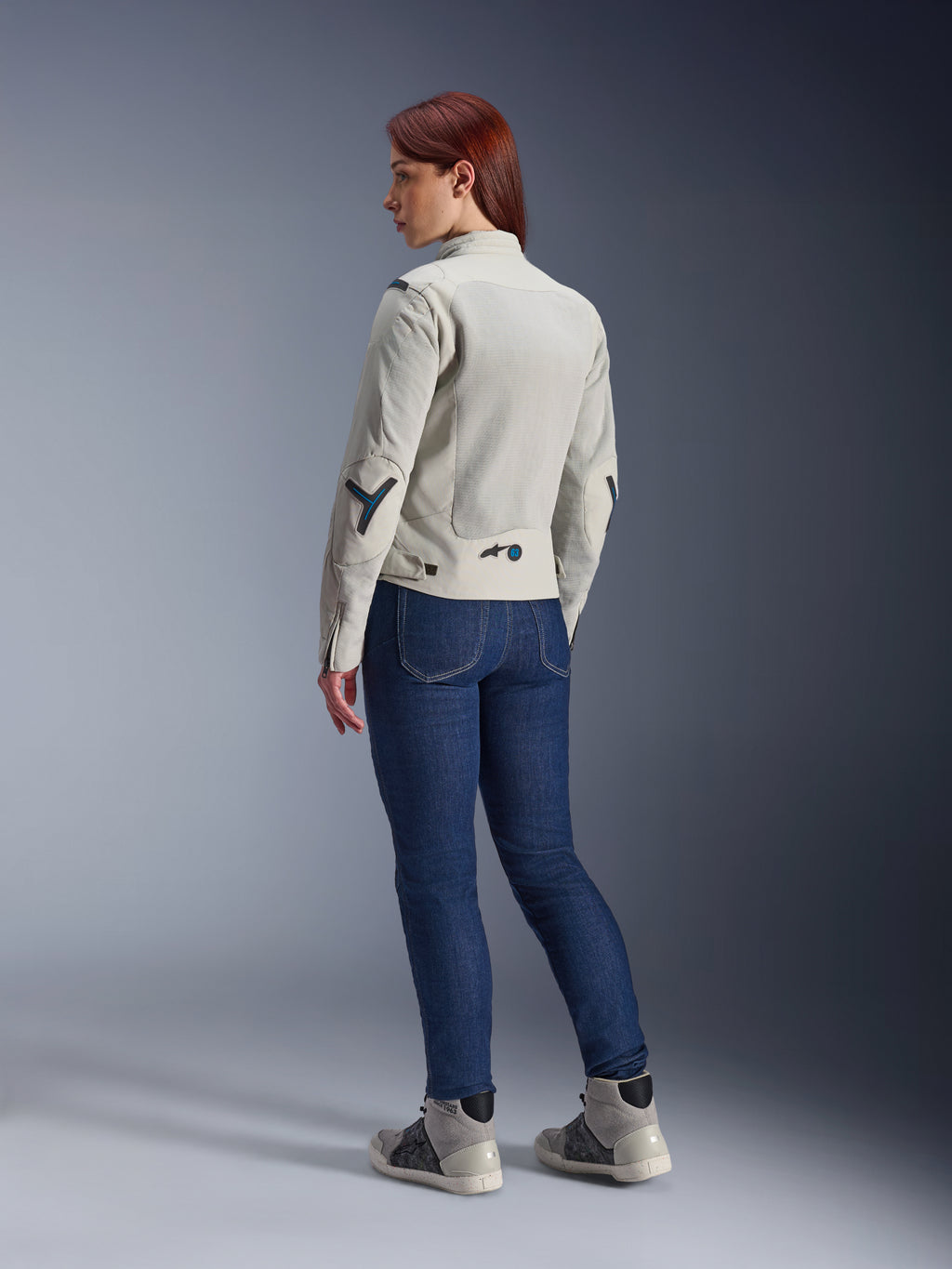 Kobieta w kurtce Alpinestars Stella Circal-1 Air w kolorze Metal Gray i Methyl Blue, widok z tyłu prezentujący siatkowe panele wentylacyjne i neonowe niebieskie akcenty, zestawiona z ciemnoniebieskimi dżinsami motocyklowymi i szarymi sneakersami, ujęcie w miejskim stylu motocyklowym.