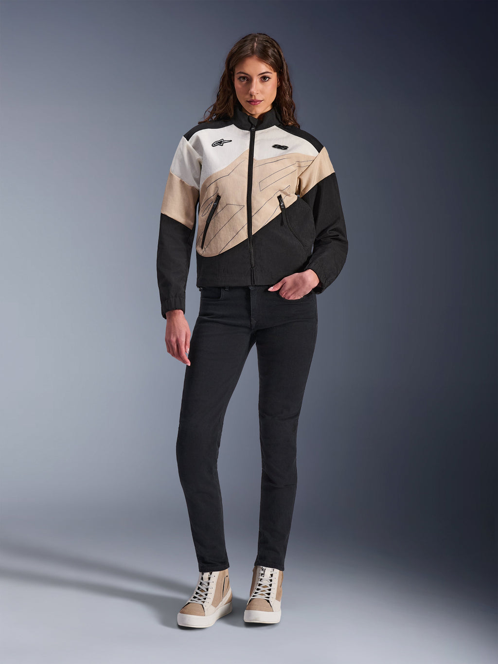 Alpinestars Woman Stella Moshell Jacket, miejska kurtka motocyklowa o asymetrycznym wzorze blokowym w kolorach czarnym, piaskowym i złamanej bieli, noszona z czarnymi dopasowanymi spodniami i beżowymi wysokimi trampkami, nowoczesna estetyka streetwear dla motocyklistek.