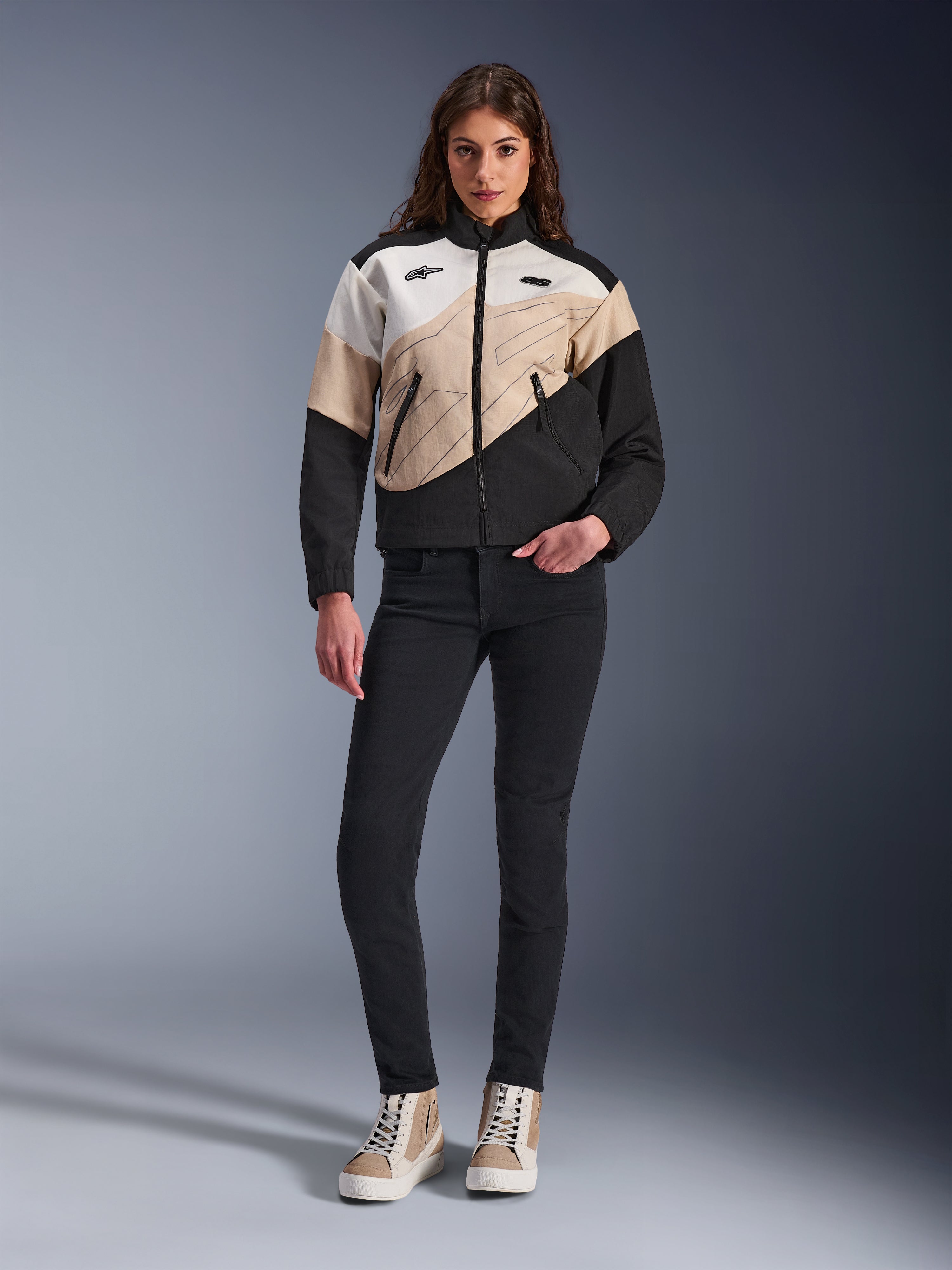 Alpinestars Woman Stella Moshell Jacket, miejska kurtka motocyklowa o asymetrycznym wzorze blokowym w kolorach czarnym, piaskowym i złamanej bieli, noszona z czarnymi dopasowanymi spodniami i beżowymi wysokimi trampkami, nowoczesna estetyka streetwear dla motocyklistek.
