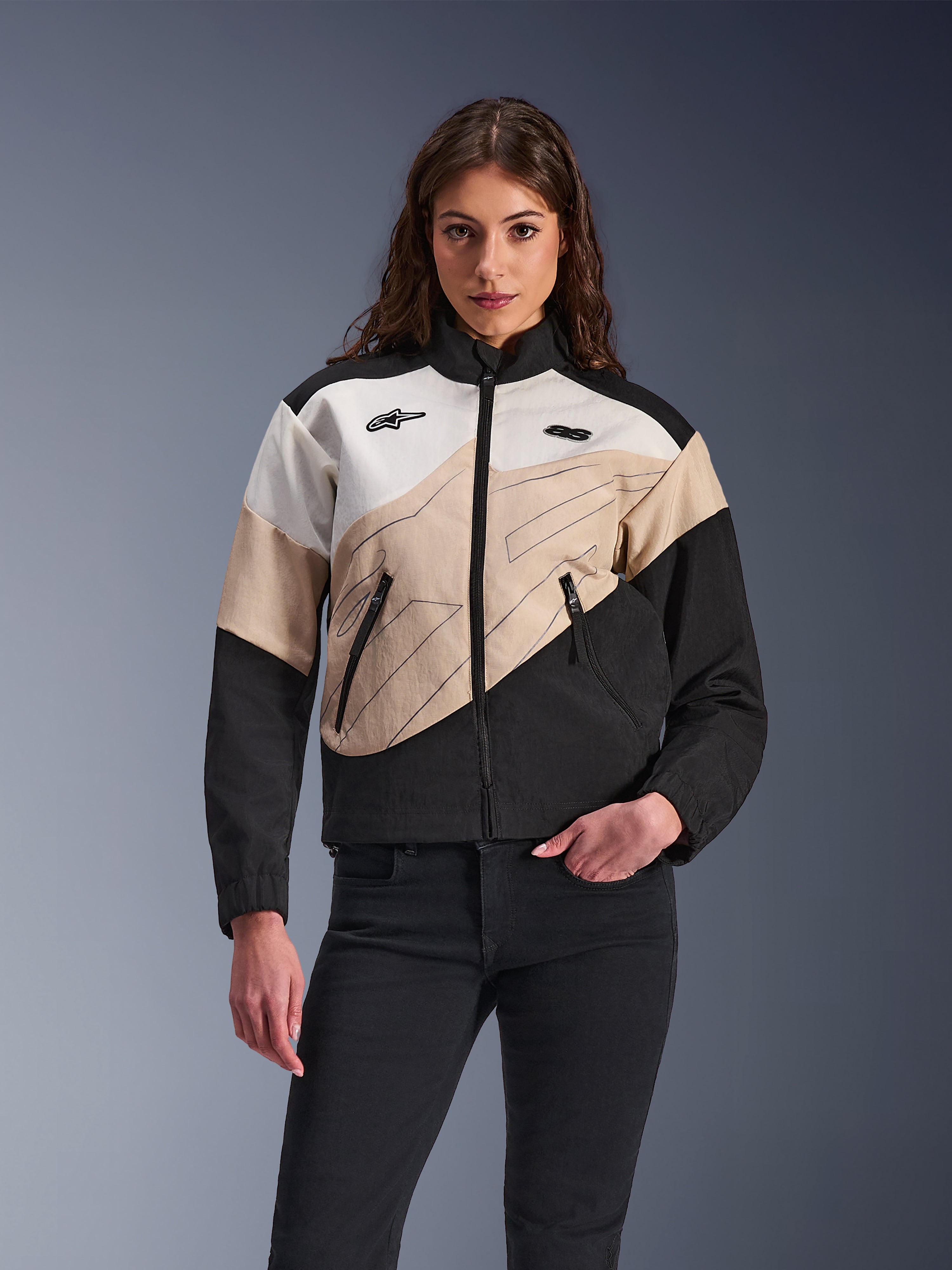 Alpinestars Stella Moshell Jacket, damska miejska kurtka motocyklowa, asymetryczny design typu color-blocking w kolorach czarnym, piaskowym i złamanej bieli, przedni zamek błyskawiczny i kieszenie na suwak, zestawiona z czarnymi spodniami w nowoczesnym stylu streetwear