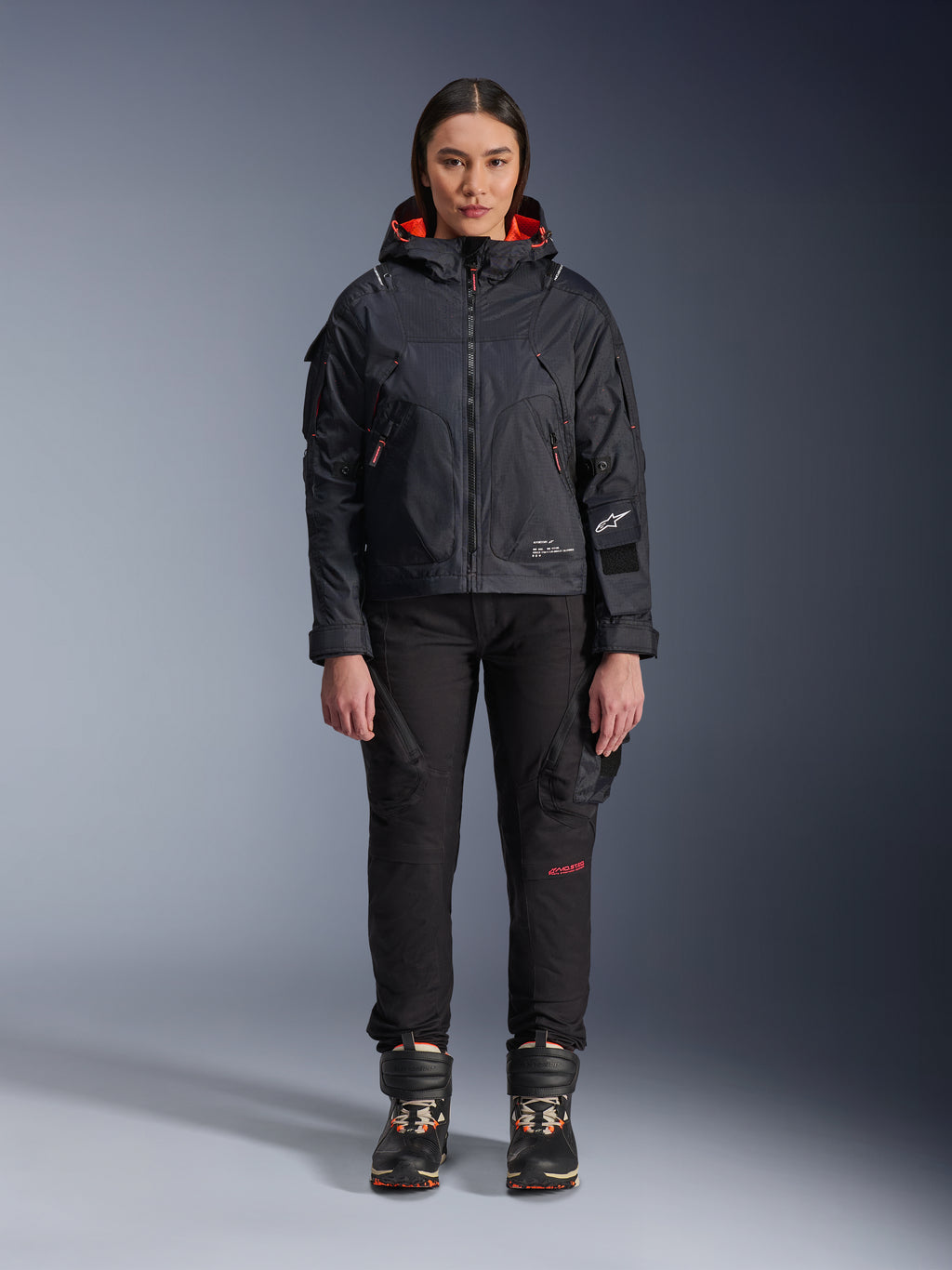 Modelka w kurtce Alpinestars Molly Air Jacket w kolorze Black i Fluo Coral, o designie inspirowanym stylem użytkowym z kapturem i koralową podszewką, w zestawieniu z czarnymi spodniami i butami motocyklowymi, współczesna miejska odzież motocyklowa