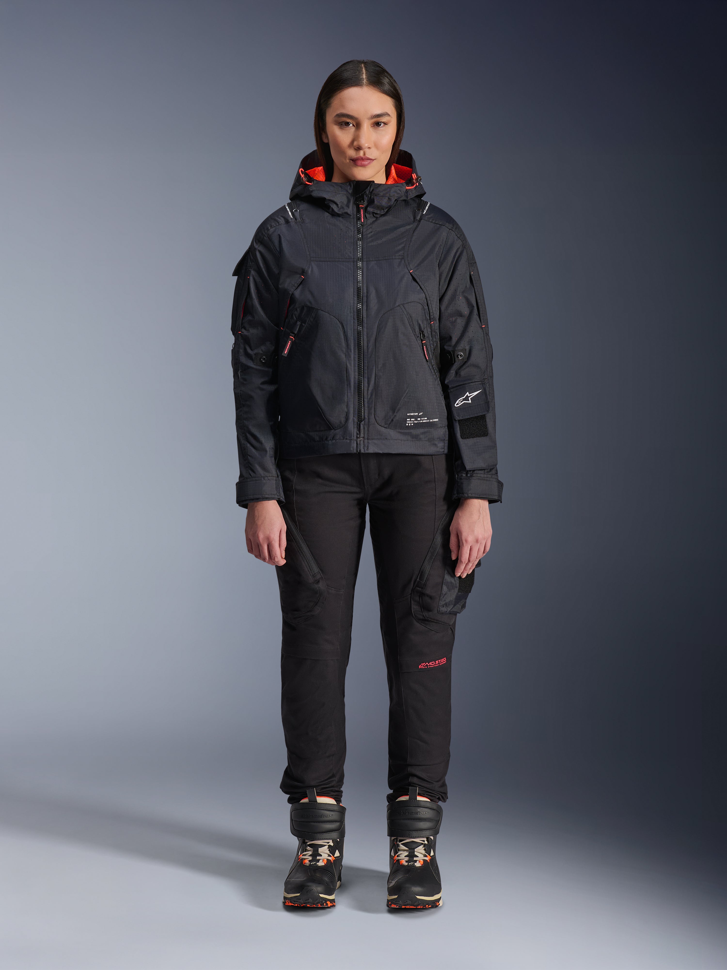 Modelka w kurtce Alpinestars Molly Air Jacket w kolorze Black i Fluo Coral, o designie inspirowanym stylem użytkowym z kapturem i koralową podszewką, w zestawieniu z czarnymi spodniami i butami motocyklowymi, współczesna miejska odzież motocyklowa