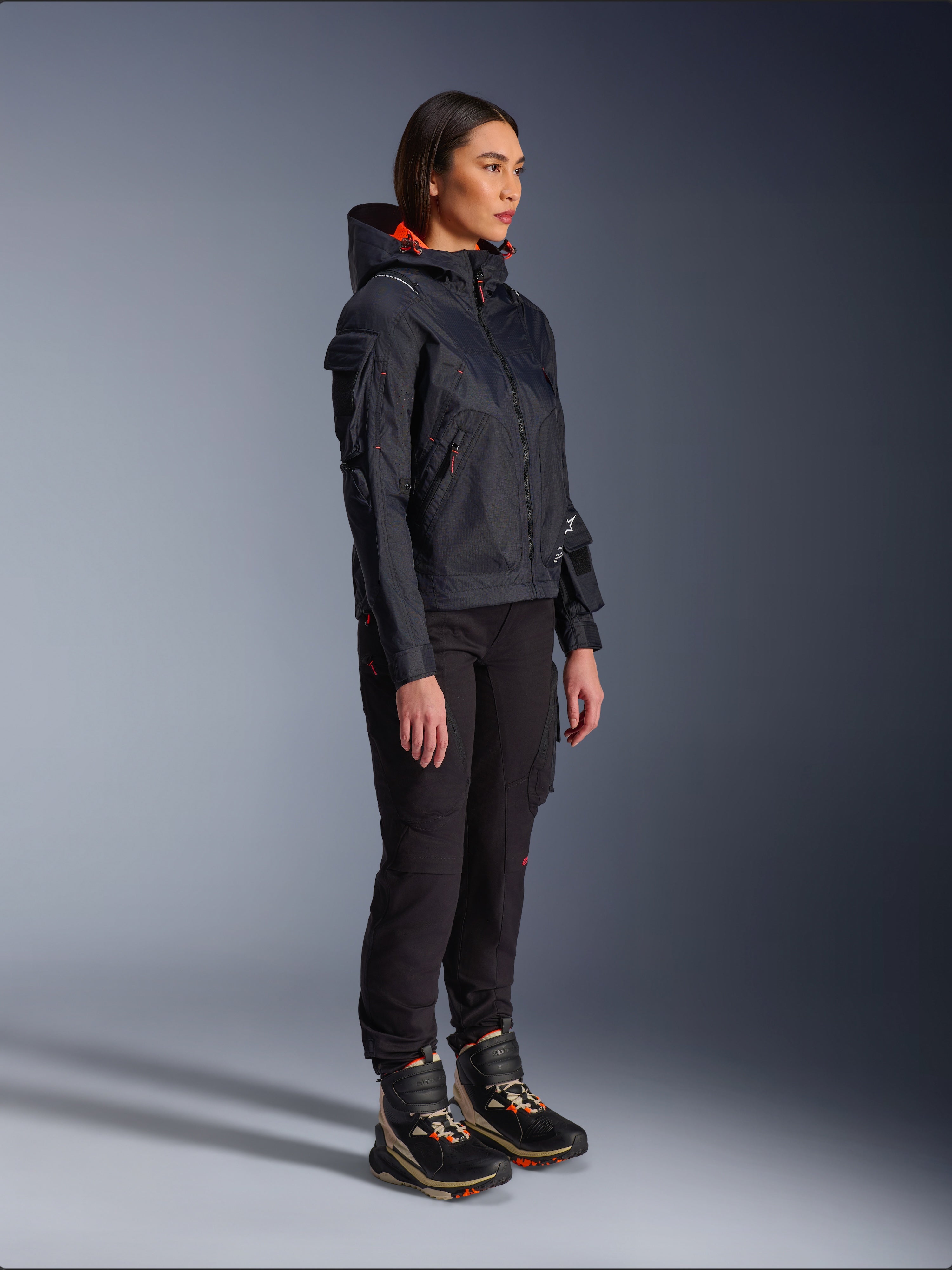 Motocyklistka ubrana w kurtkę Alpinestars Molly Air Jacket w kolorze Black Fluo Coral, lekka tekstylna kurtka motocyklowa z jaskrawopomarańczową podszewką kaptura, zestawiona z czarnymi spodniami motocyklowymi i butami technicznymi, nowoczesny miejski design inspirowany stylem utility.