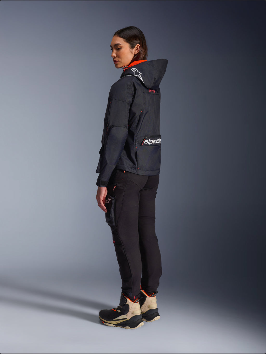 Alpinestars Actea Women's Pants, czarne, nowoczesne spodnie motocyklowe o zwężanym kroju i z kieszenią cargo na udzie, widok z boku i od tyłu na modelce ubranej w ciemną kurtkę z kapturem i buty motocyklowe, miejski styl jazdy.
