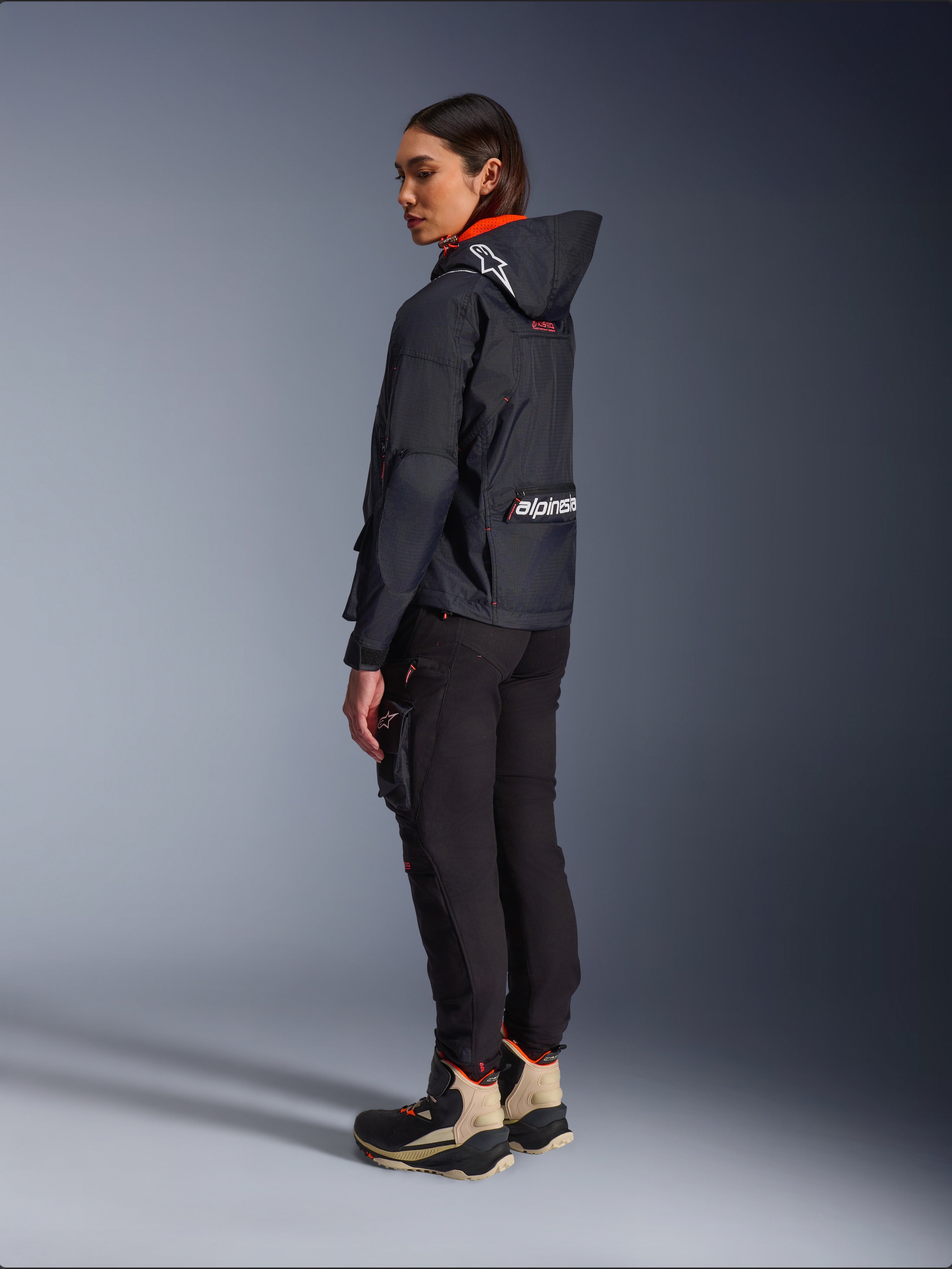 Alpinestars Actea Women's Pants, czarne, nowoczesne spodnie motocyklowe o zwężanym kroju i z kieszenią cargo na udzie, widok z boku i od tyłu na modelce ubranej w ciemną kurtkę z kapturem i buty motocyklowe, miejski styl jazdy.