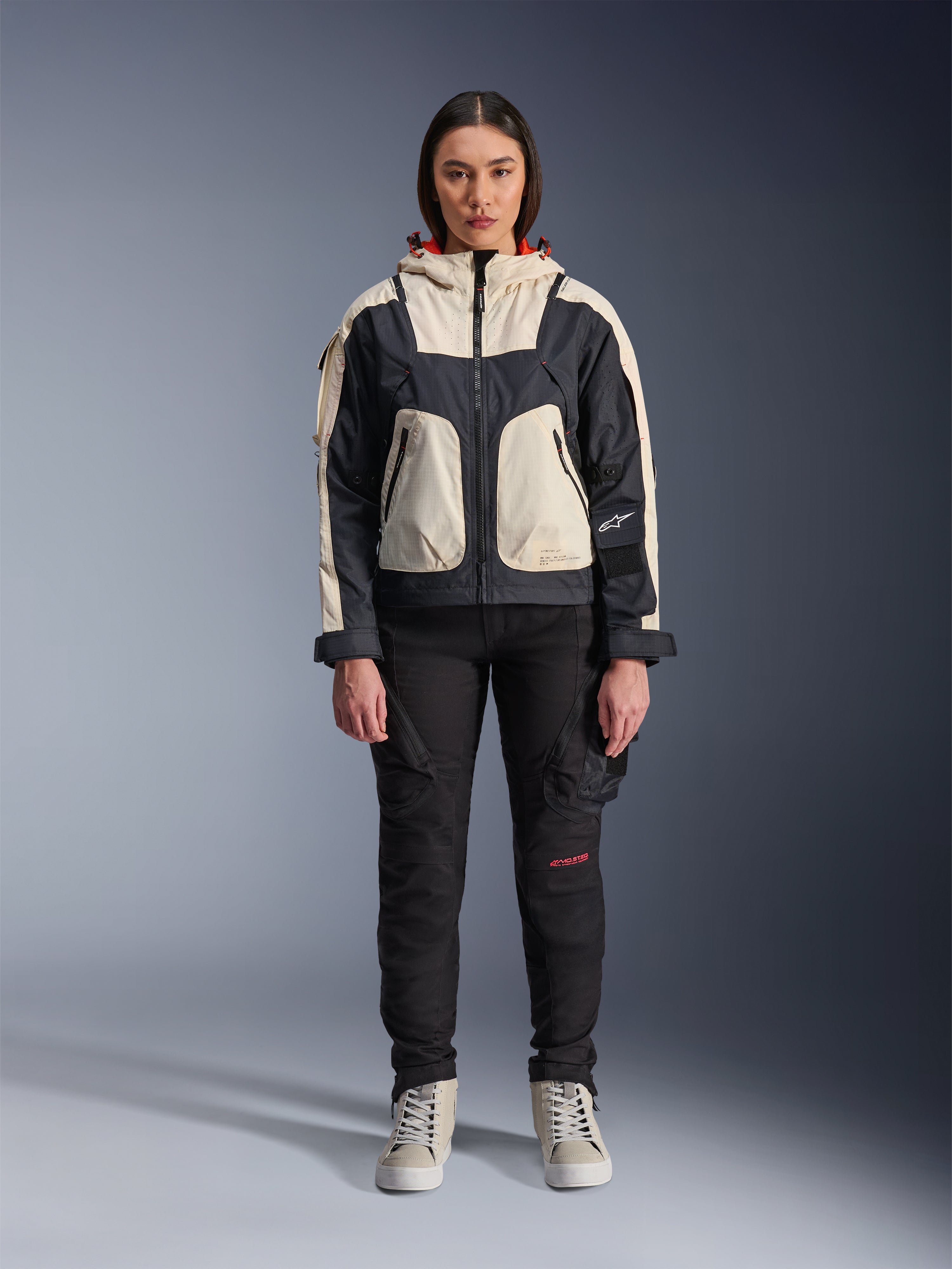 Modelka w kurtce Alpinestars Molly Air Jacket w kolorze Black Almond, design typu color-block z kremowymi i czarnymi panelami oraz kapturem z czerwoną podszewką, czarne spodnie motocyklowe i kremowe sneakersy, studyjne zdjęcie współczesnej miejskiej odzieży motocyklowej